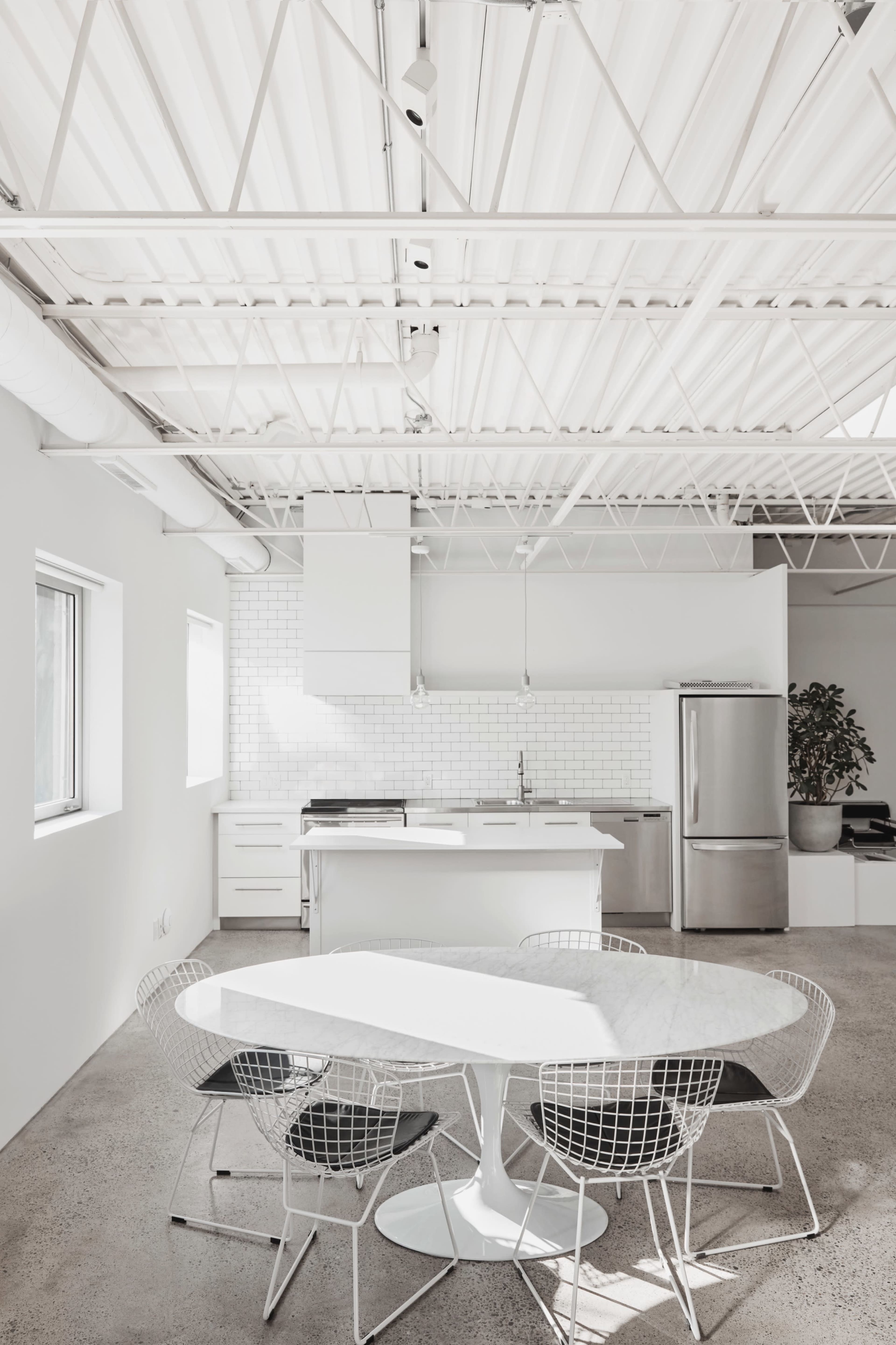 A modern kitchen features a white dining table surrounded by wire chairs and stainless steel appliances in a bright, open space.