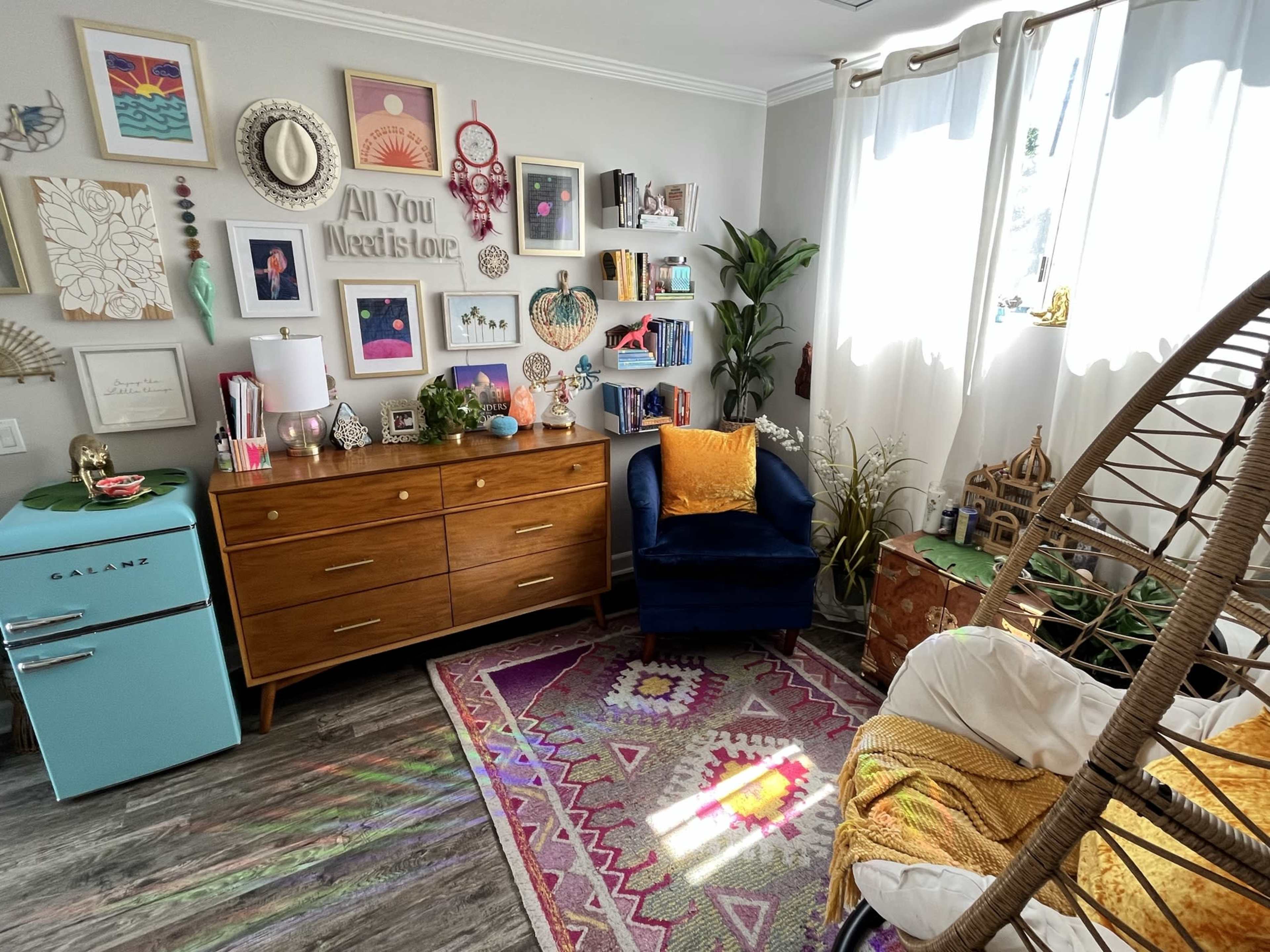 A cozy room featuring a blue chair, a wooden dresser, and decorated walls with various framed pictures and shelves filled with books and decorative items.
