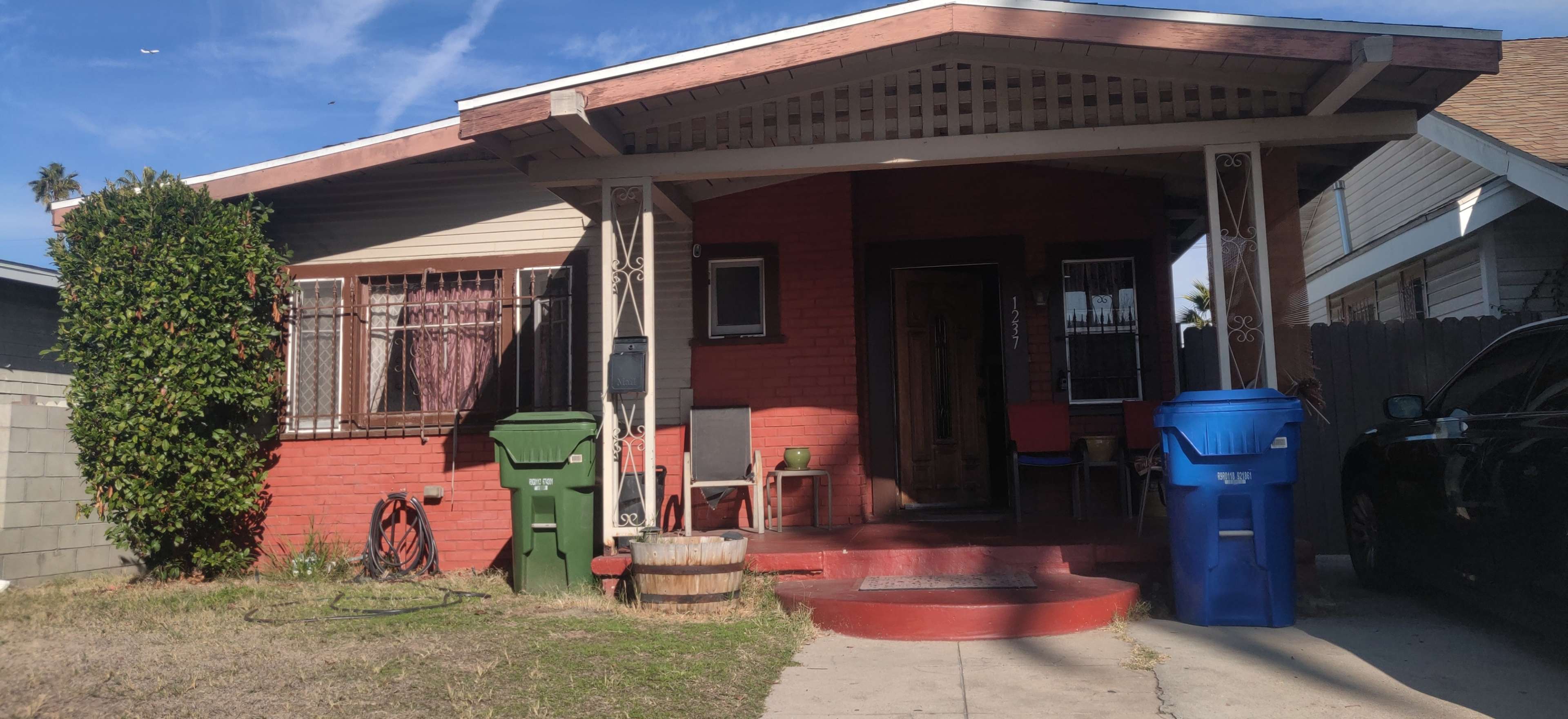 The image shows a single-story house with a red brick façade, a porch, and two trash bins in front, one green and one blue.