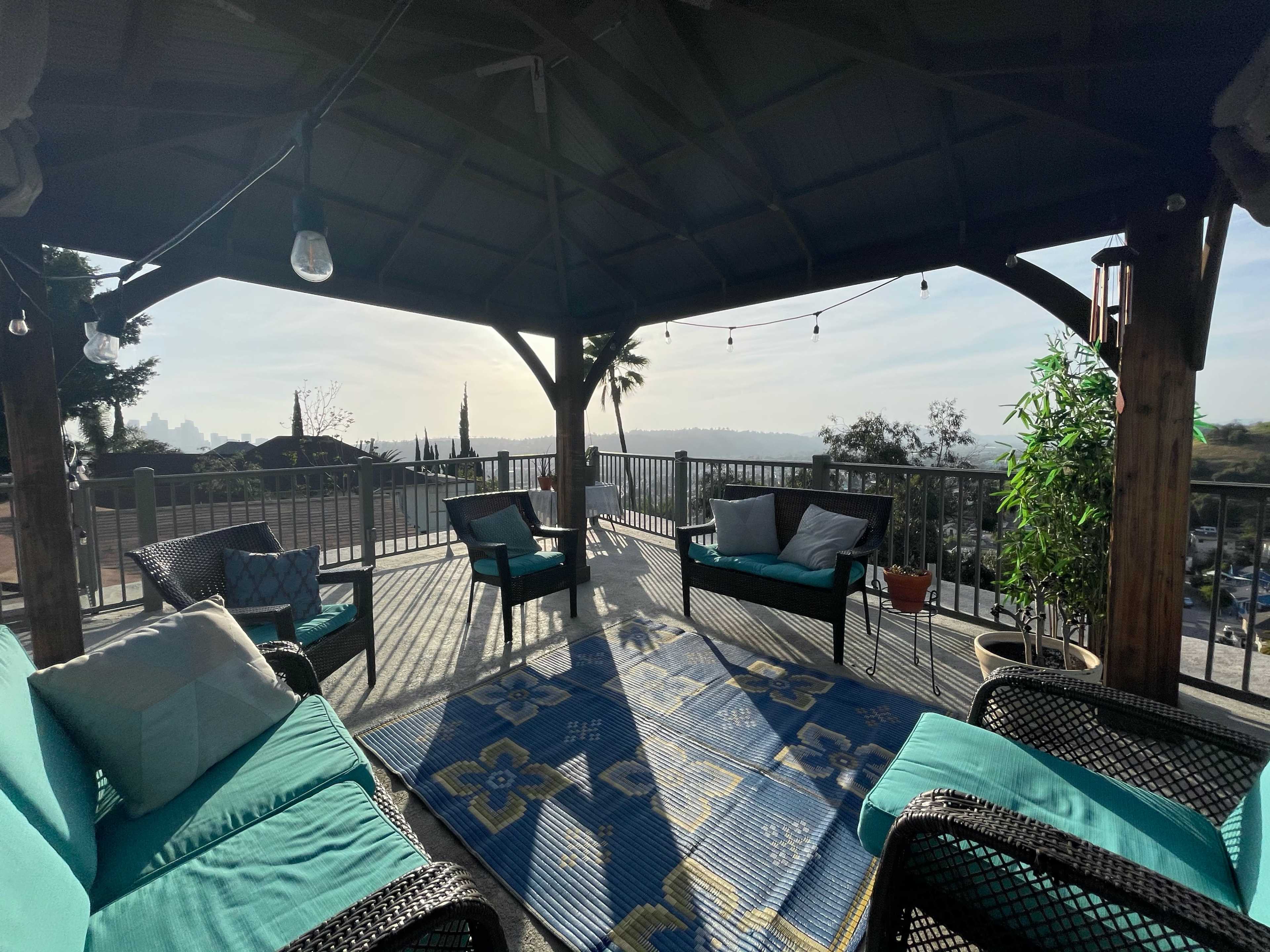 A covered outdoor seating area features wicker chairs with teal cushions, a large area rug, and a view of the landscape in the distance.