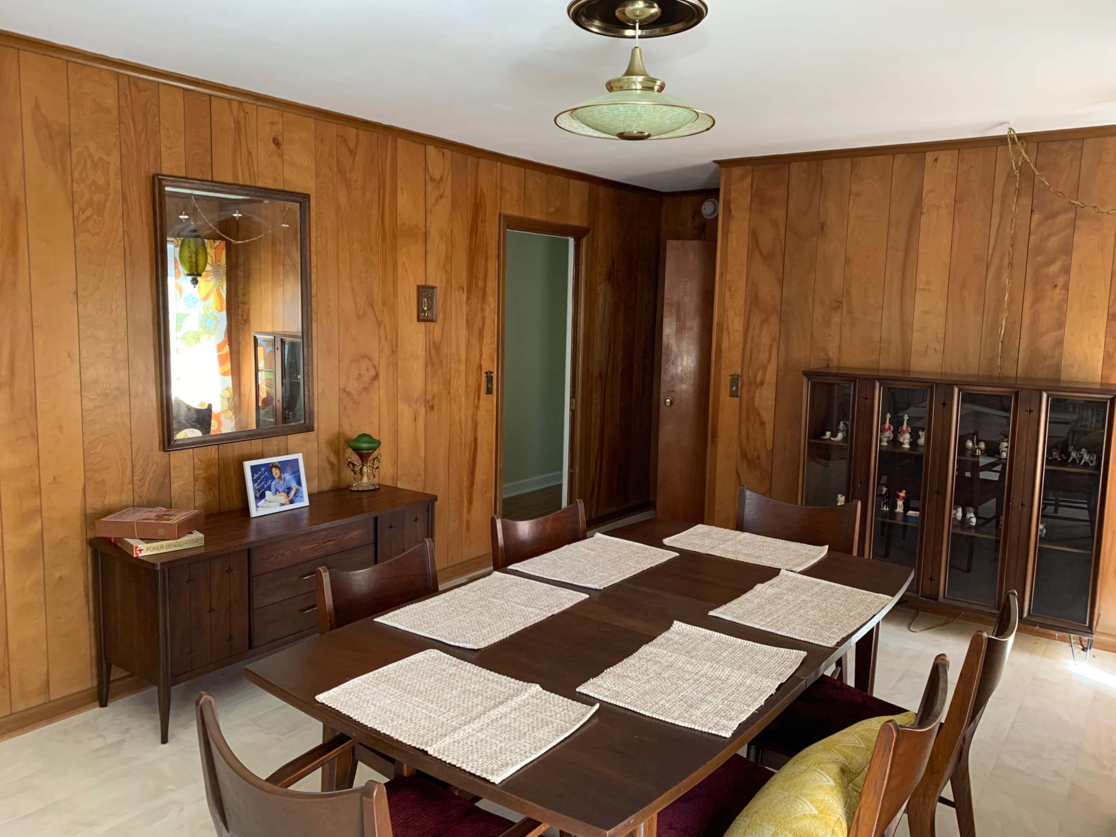 A dining room features a wooden table set with placemats, surrounded by chairs, with wood-paneled walls and a cabinet displaying various items.