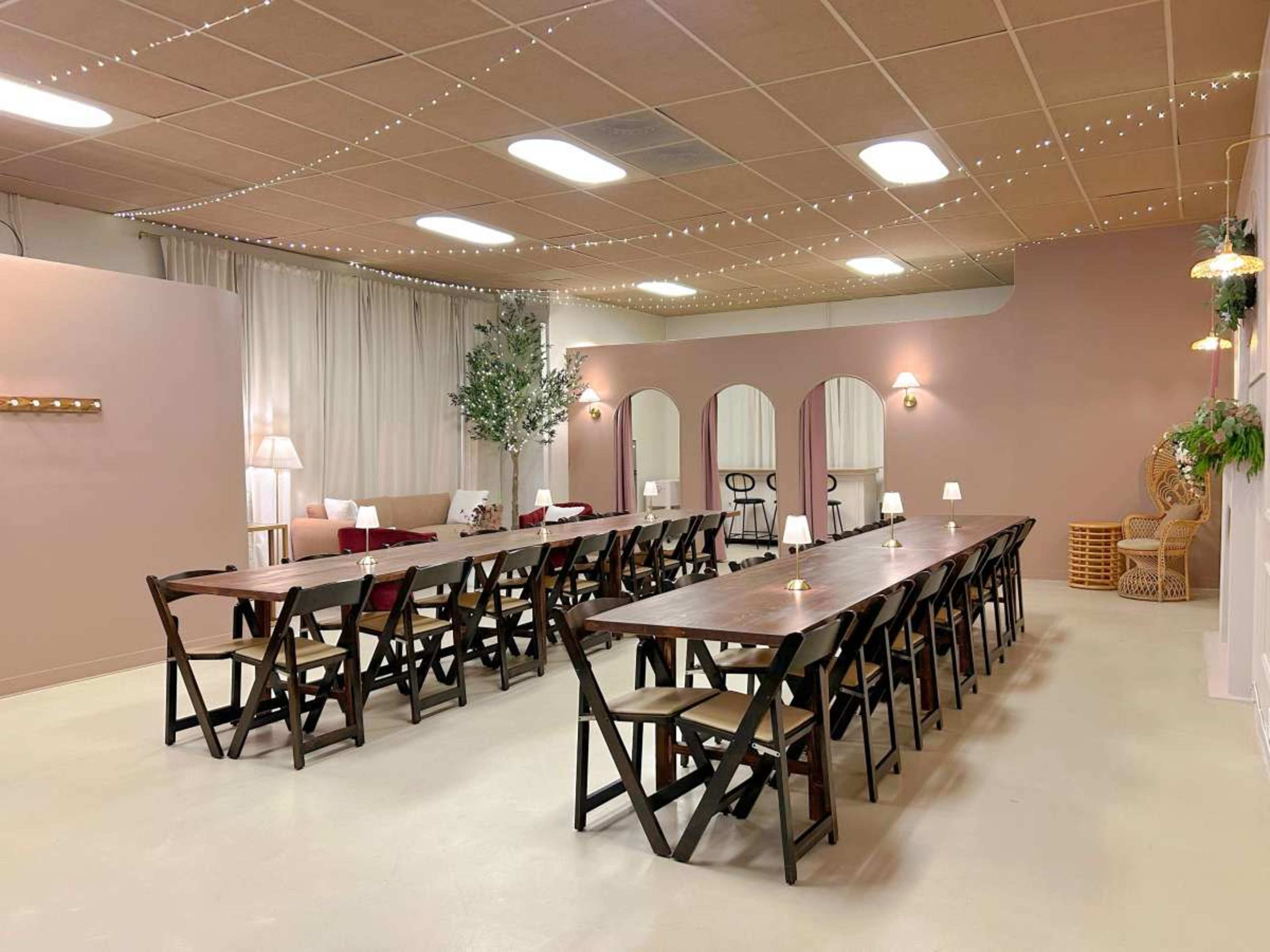 The image shows a spacious room with a long arrangement of wooden tables and chairs, accented by soft lighting and decorative plants.