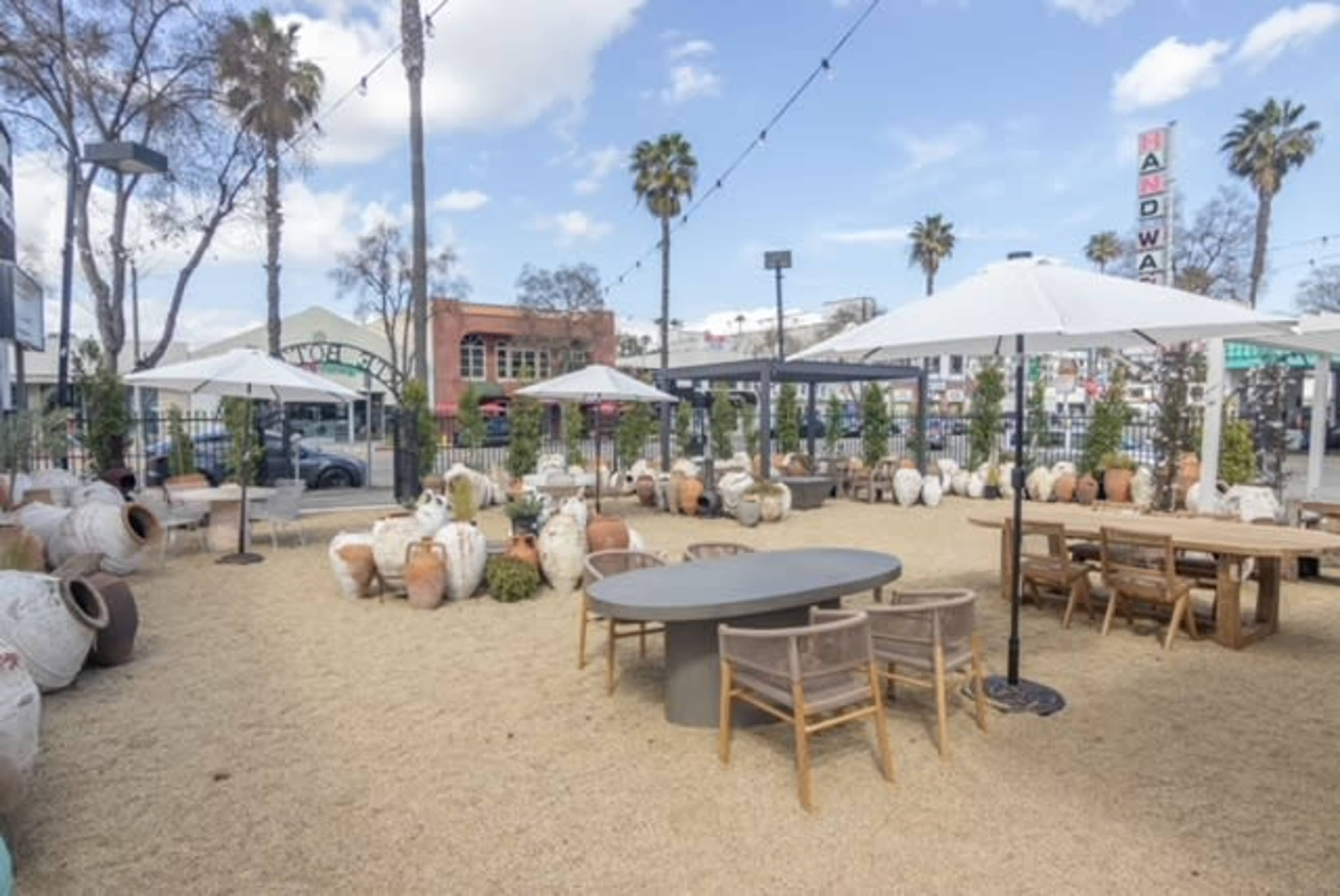 A spacious outdoor dining area features tables, chairs, and large decorative pots, surrounded by palm trees and string lights against a backdrop of buildings.