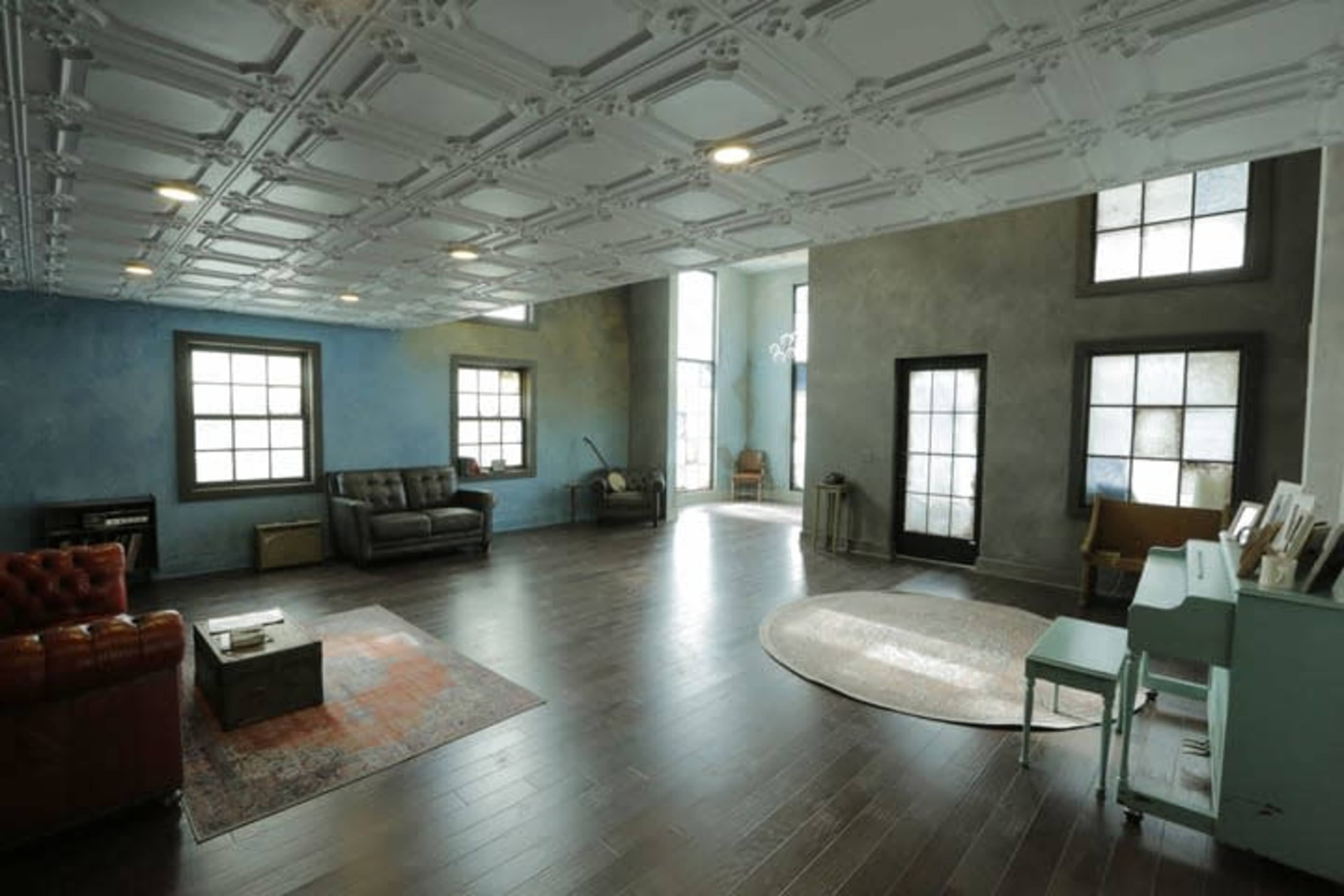 The image shows a spacious interior room with hardwood floors, large windows, and a mix of modern and vintage furniture.