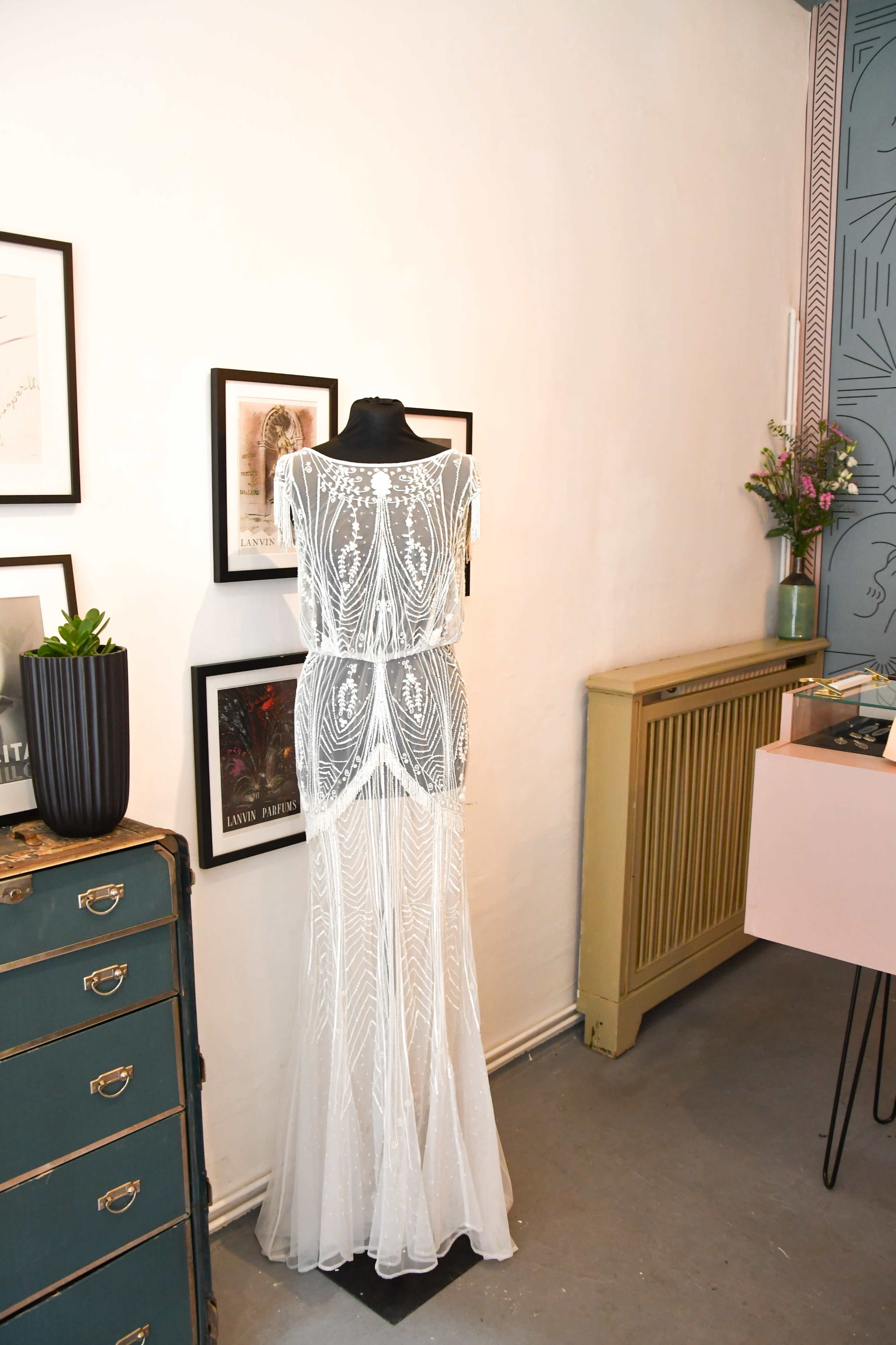 A sheer, beaded white dress is displayed on a mannequin against a backdrop of framed art and a potted plant in a stylish interior.