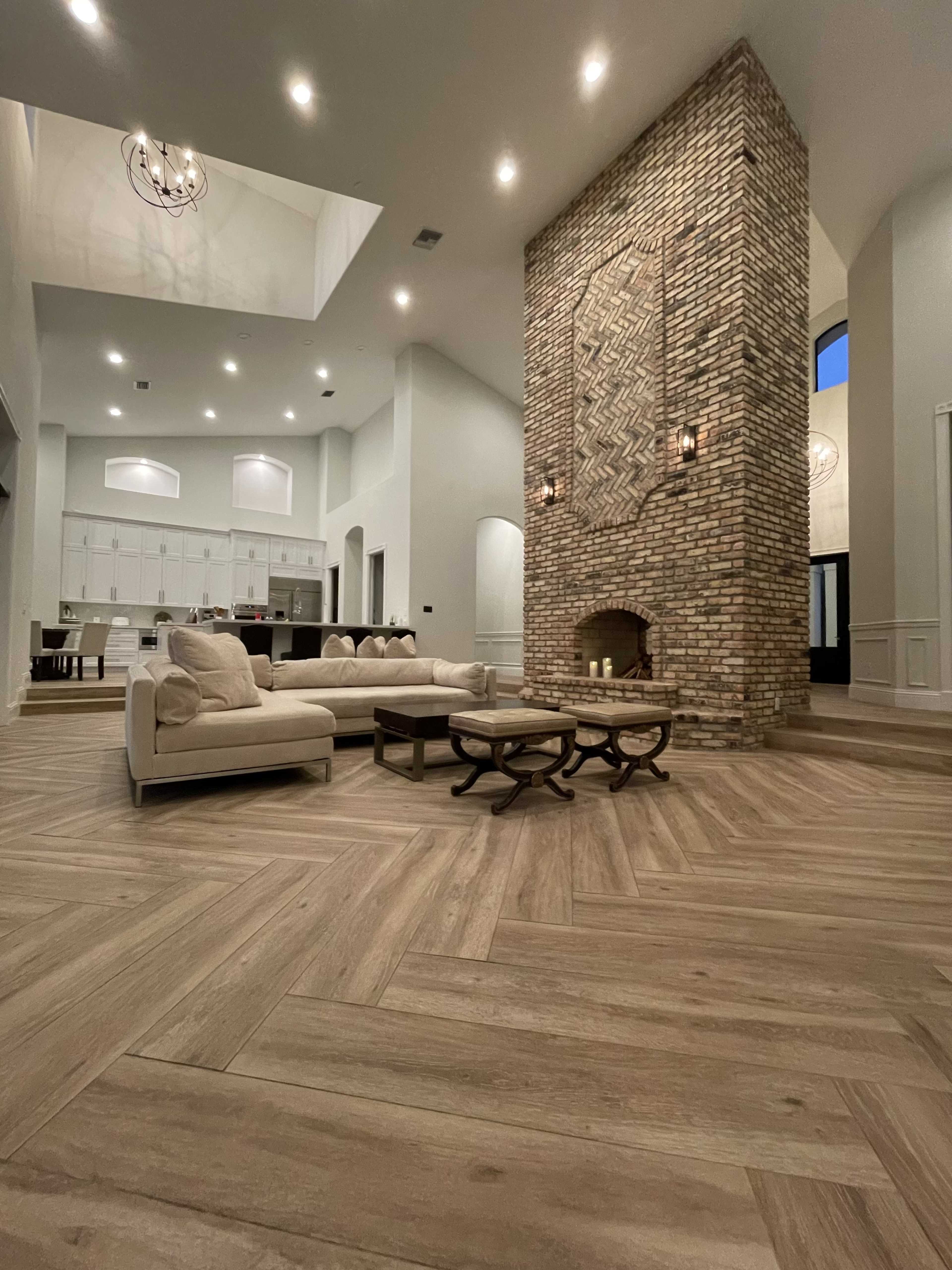A spacious living room features a tall stone fireplace and a herringbone-patterned wooden floor, with a large sectional sofa and two coffee tables arranged in the center.