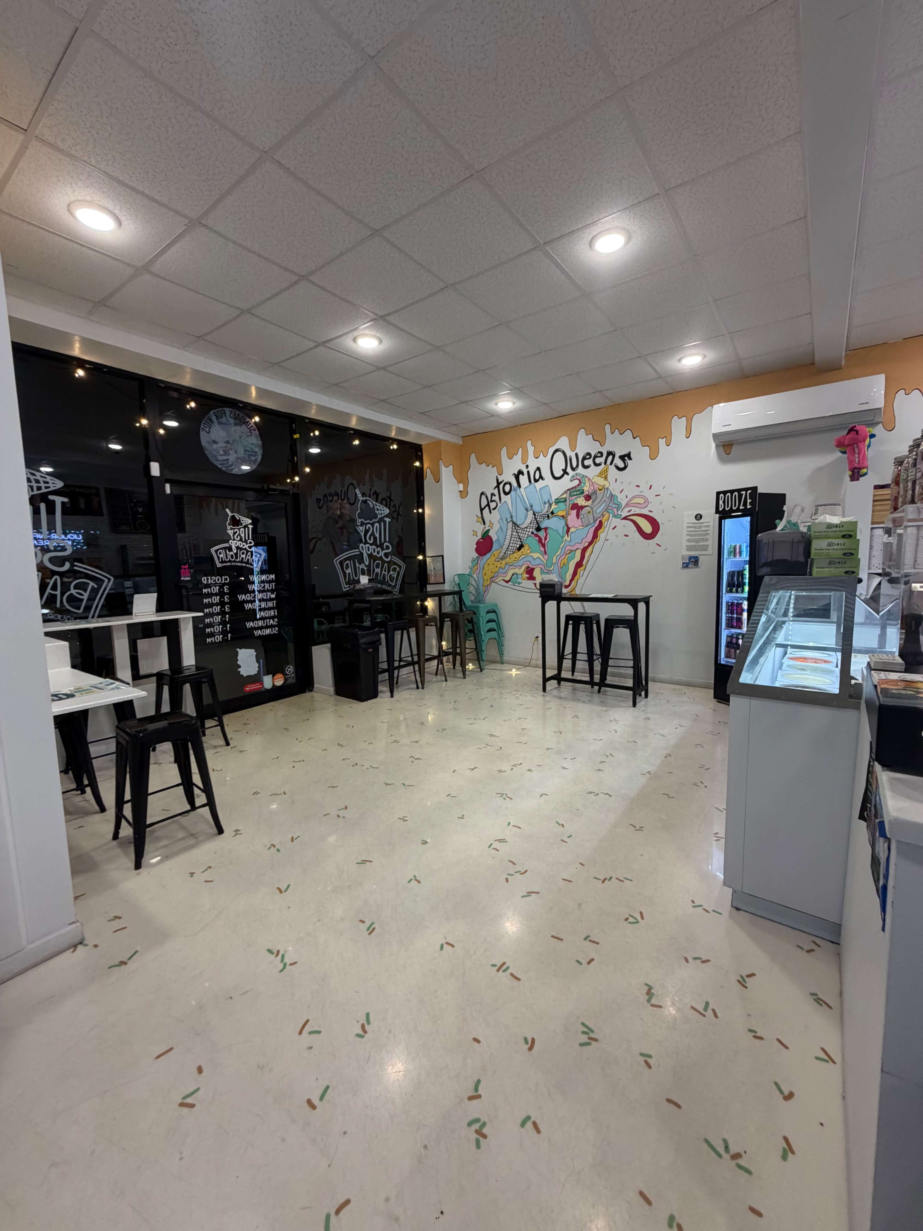 The image shows an empty ice cream shop with colorful wall art, tables, and a display counter.
