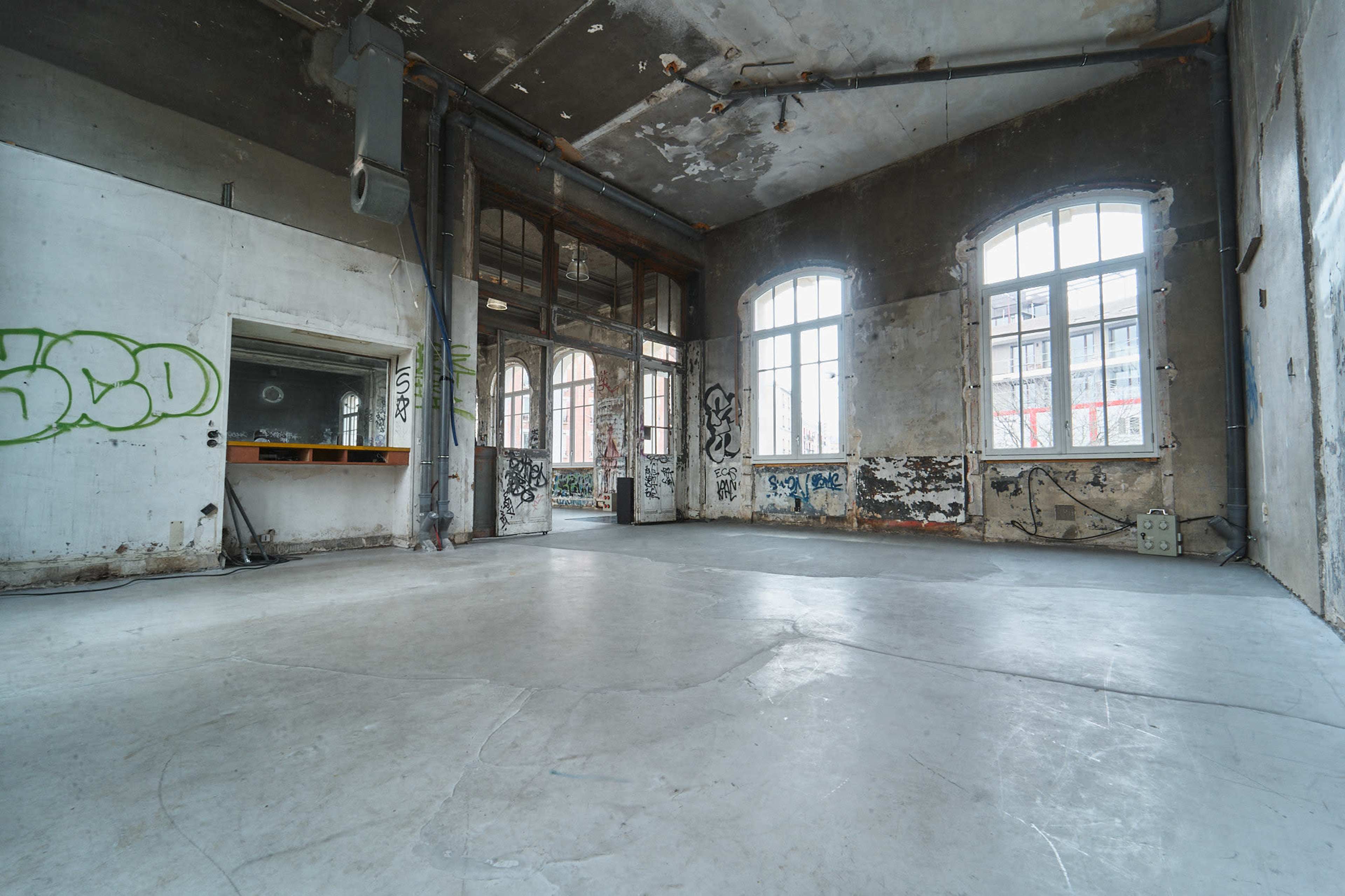 The image shows an empty, dilapidated interior space with concrete flooring, large windows, and graffiti-covered walls.