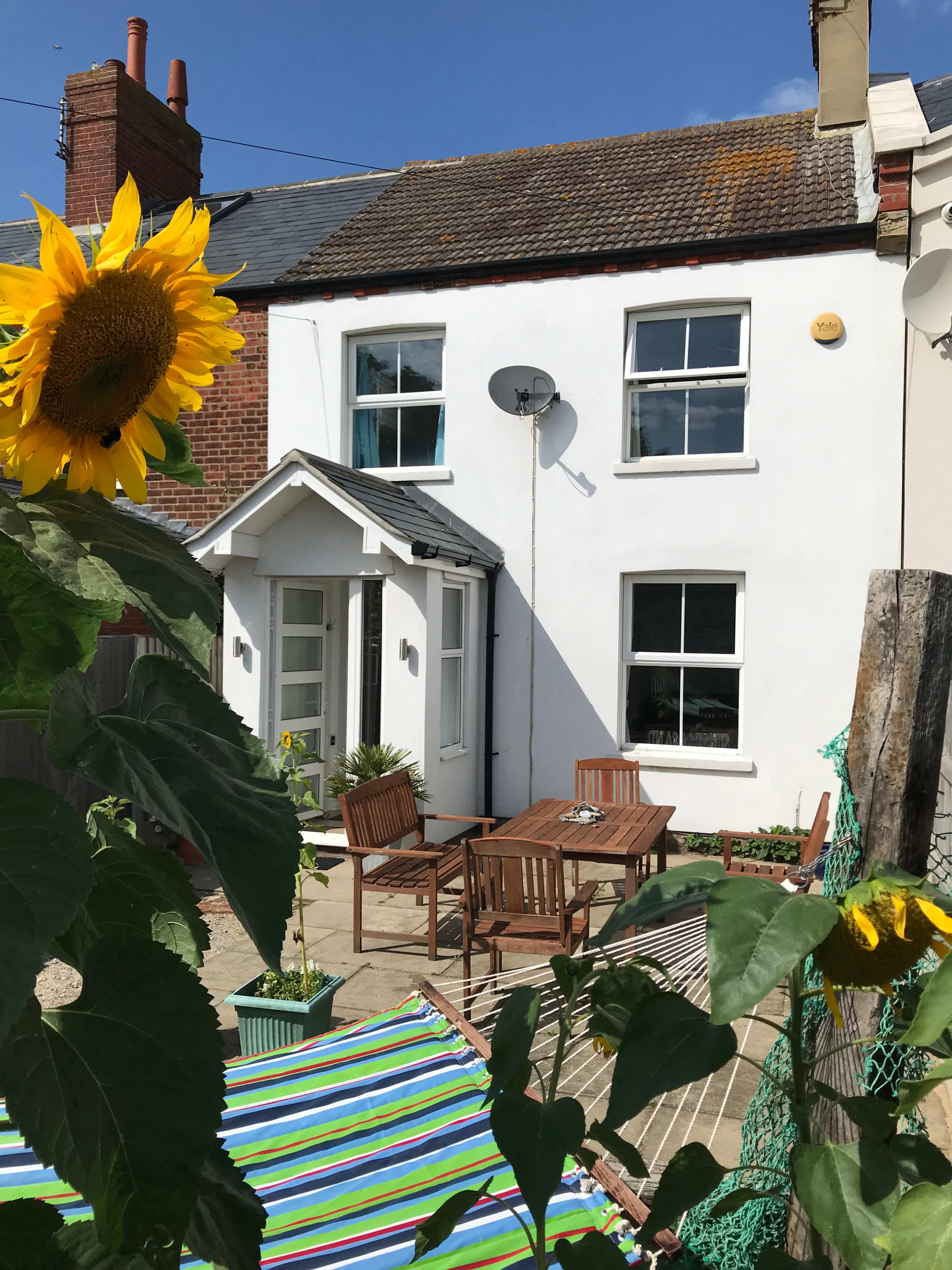 A white house with a small patio area featuring wooden furniture and a hammock, surrounded by sunflowers and greenery.