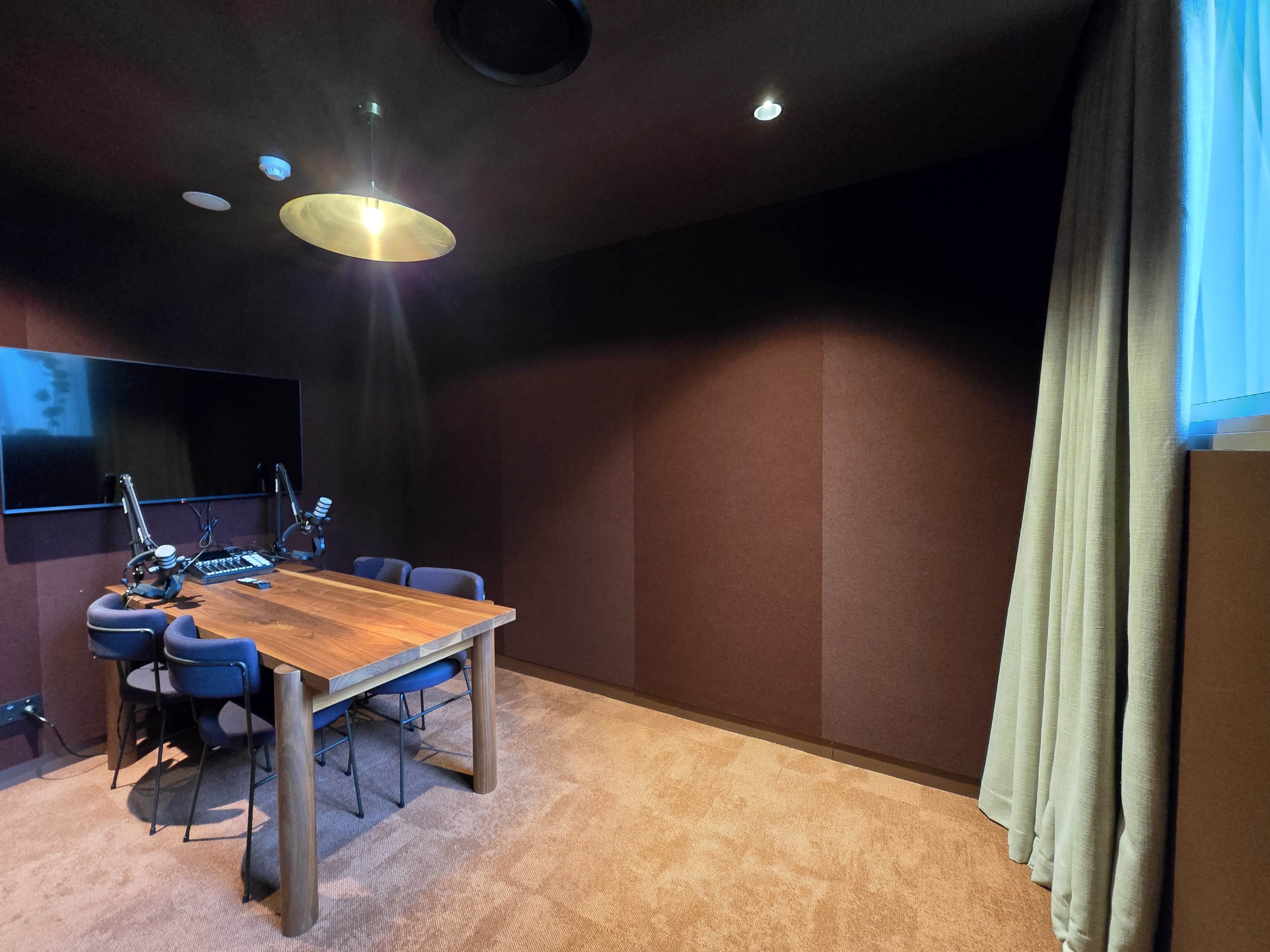 The image shows a recording studio with a wooden table, chairs, and soundproofed walls, illuminated by a ceiling light.