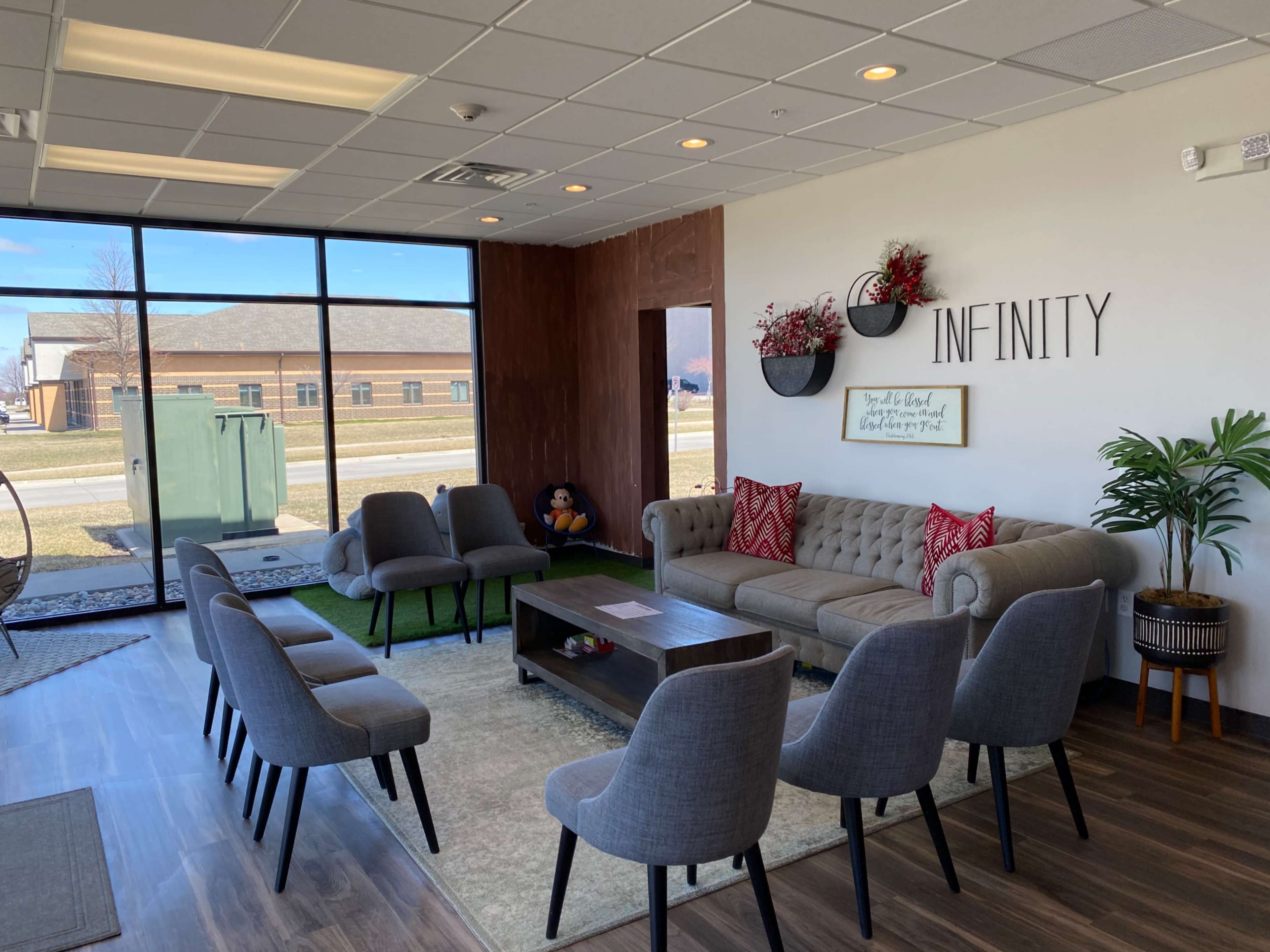 A modern waiting area with a beige sofa, a coffee table, and multiple gray chairs arranged around the room, featuring large windows that provide natural light.
