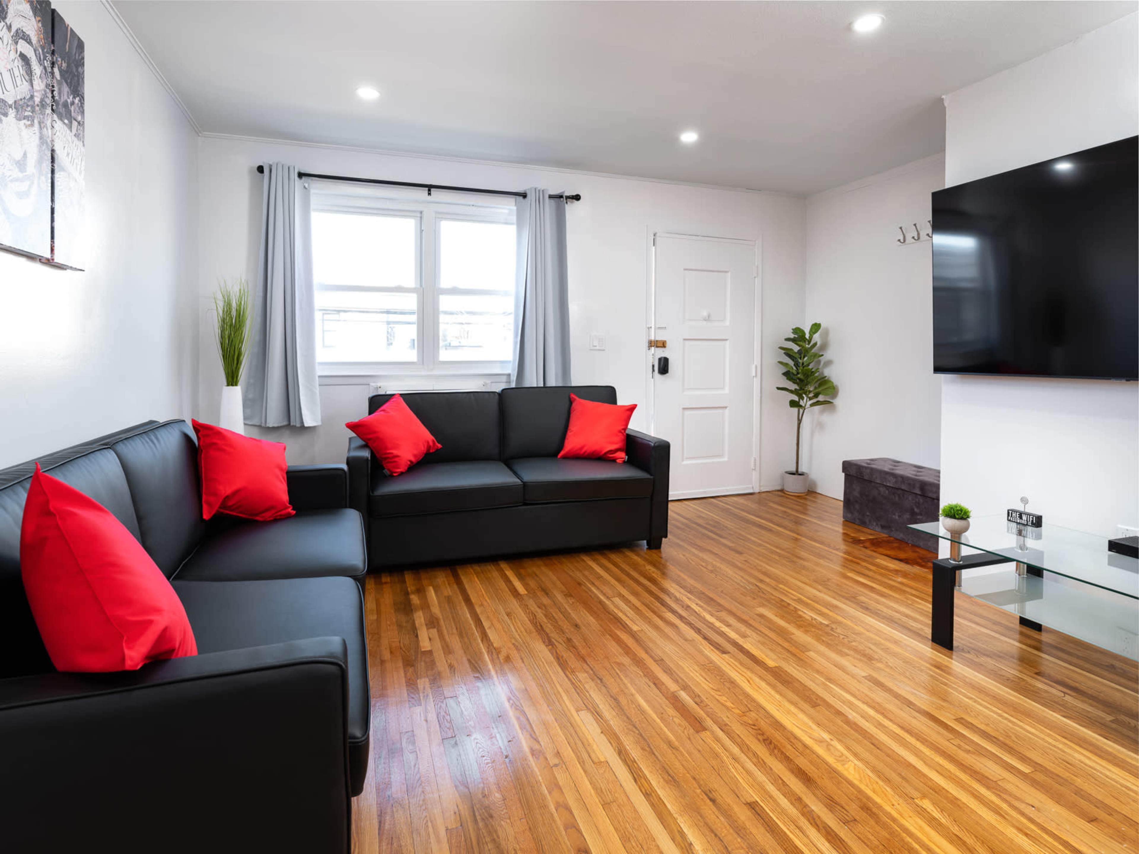 A modern living room features black sofas with red accent pillows, a glass coffee table, and a large TV mounted on the wall, all set on a wooden floor.