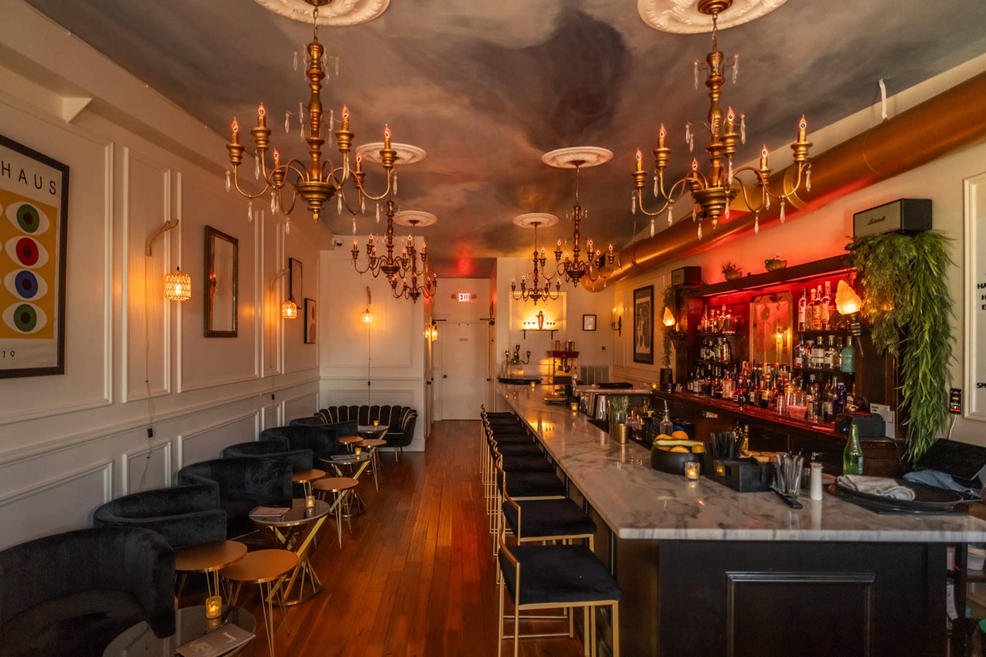 A stylish bar interior with a long marble counter, candle-lit tables, and ornate chandeliers hanging from the ceiling.