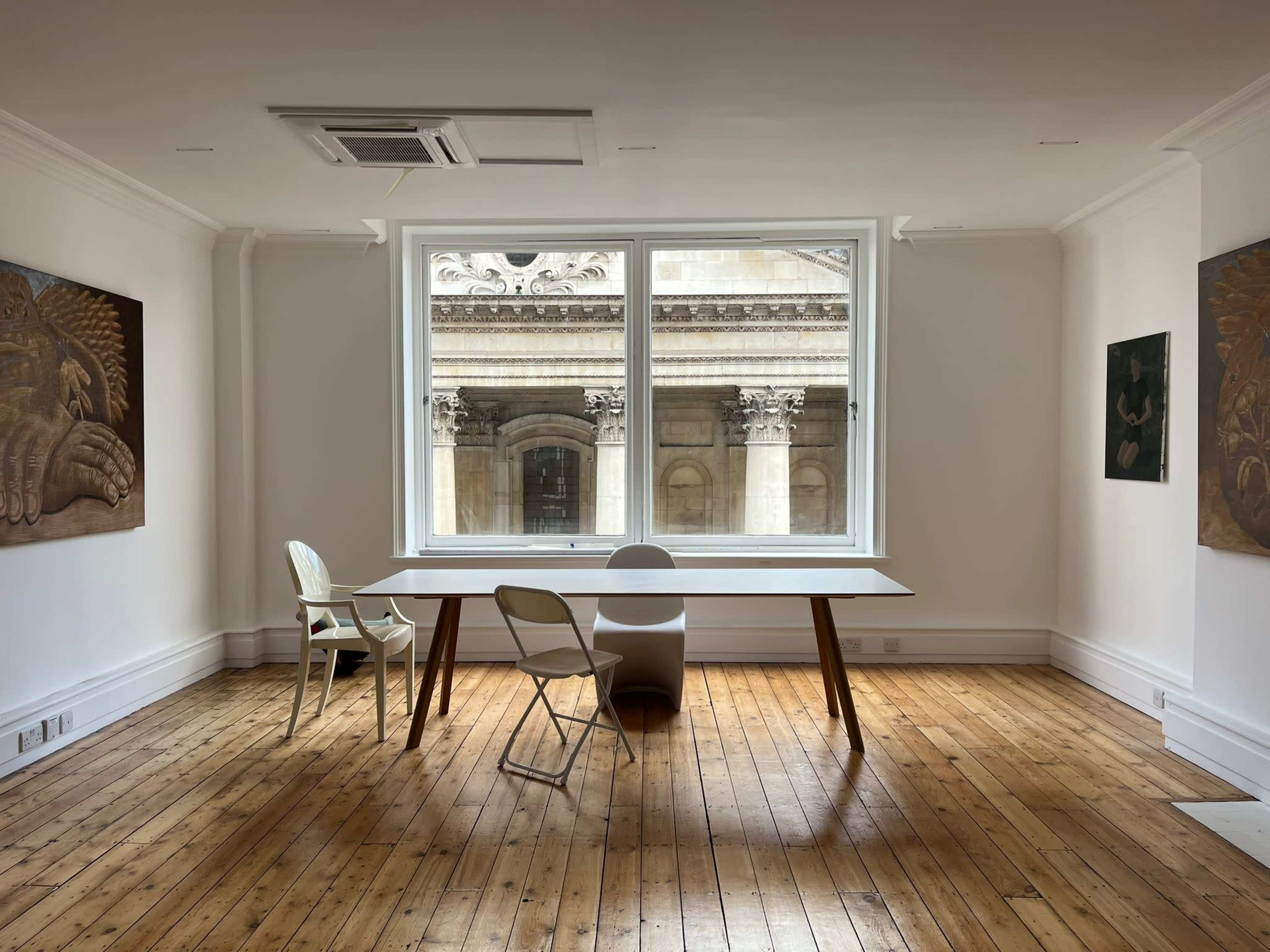 A minimalist room features a large window overlooking an ornate building, with a table and two chairs on a wooden floor.
