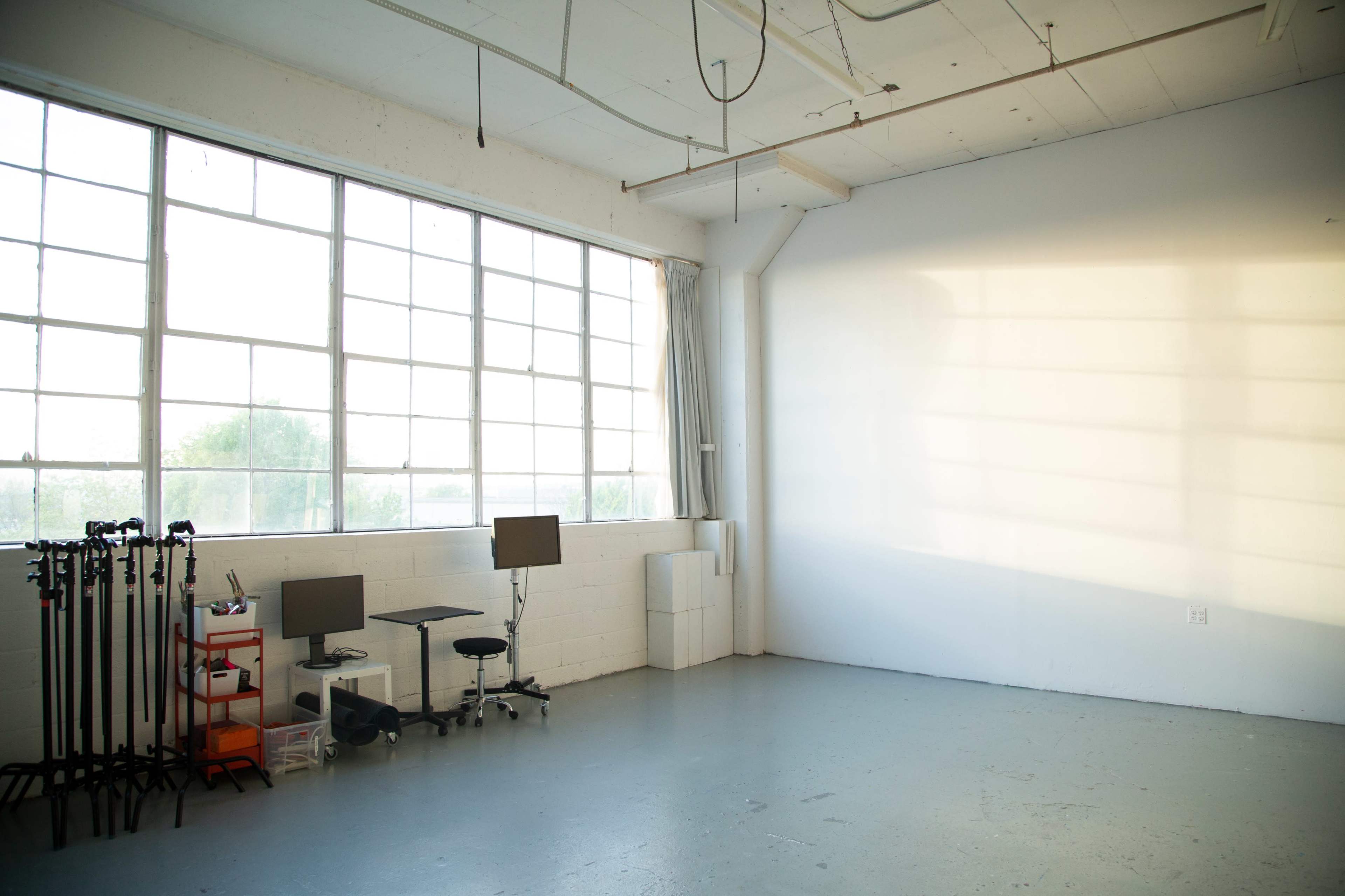 A spacious, empty room with large windows, two computer monitors, and a few pieces of furniture along one wall.
