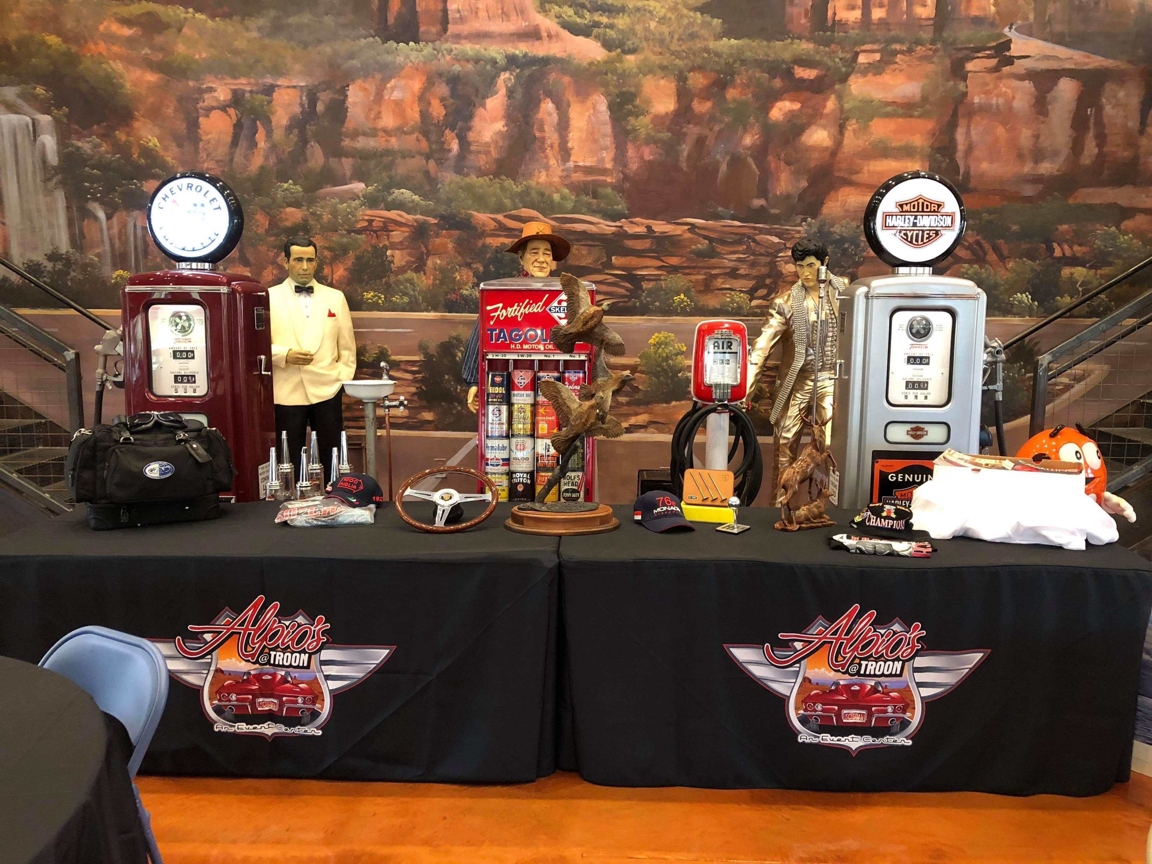 A display featuring vintage gas pumps, a mannequin dressed in a suit, a statue of a cowboy, and various nostalgic automotive memorabilia on a table with a logo banner.
