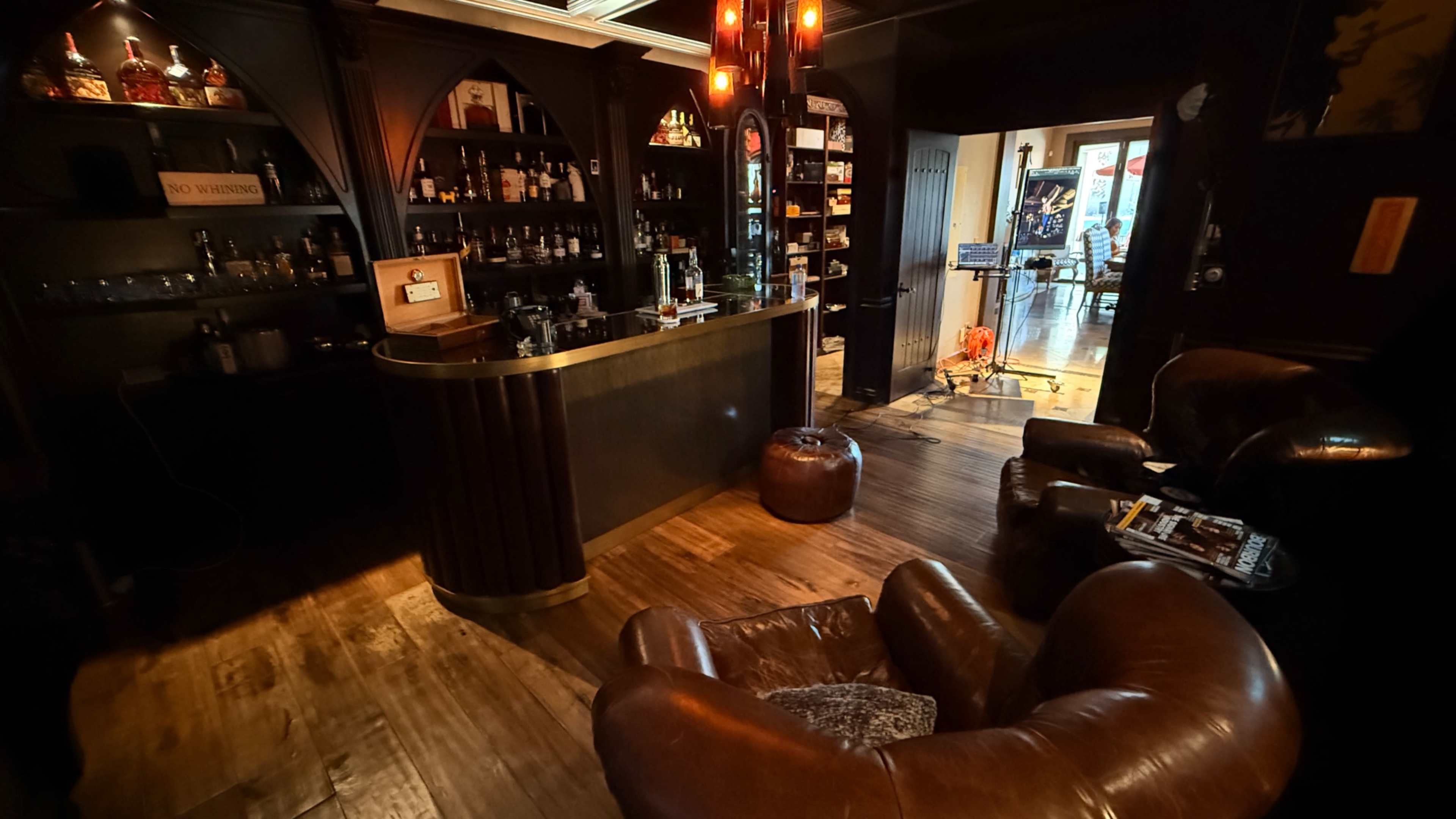A dimly lit bar area features a curved counter, shelves lined with bottles, and leather armchairs, creating a cozy seating space.