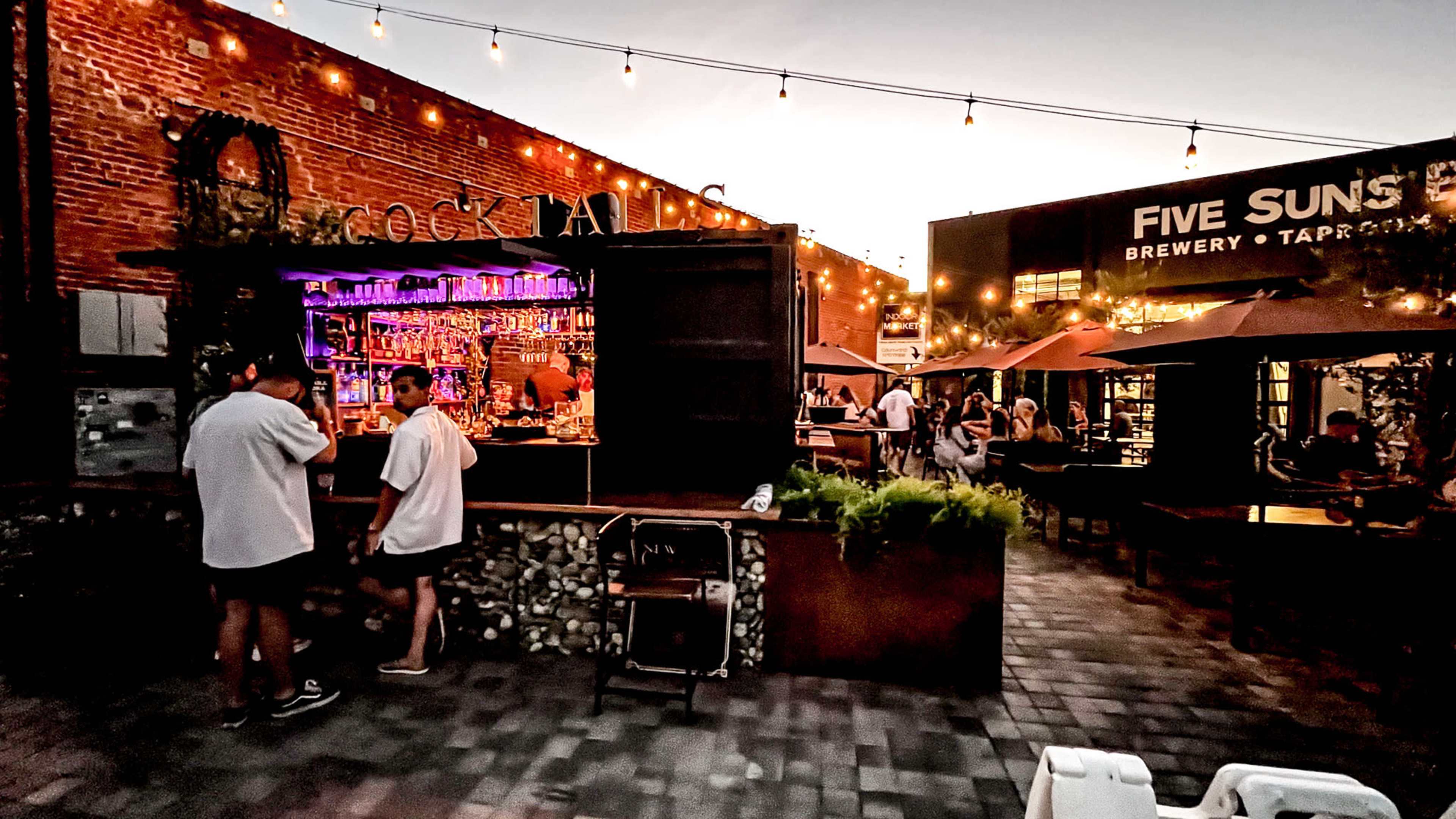 An outdoor bar scene features patrons interacting at a stylish bar illuminated by string lights, with people seated at tables nearby.