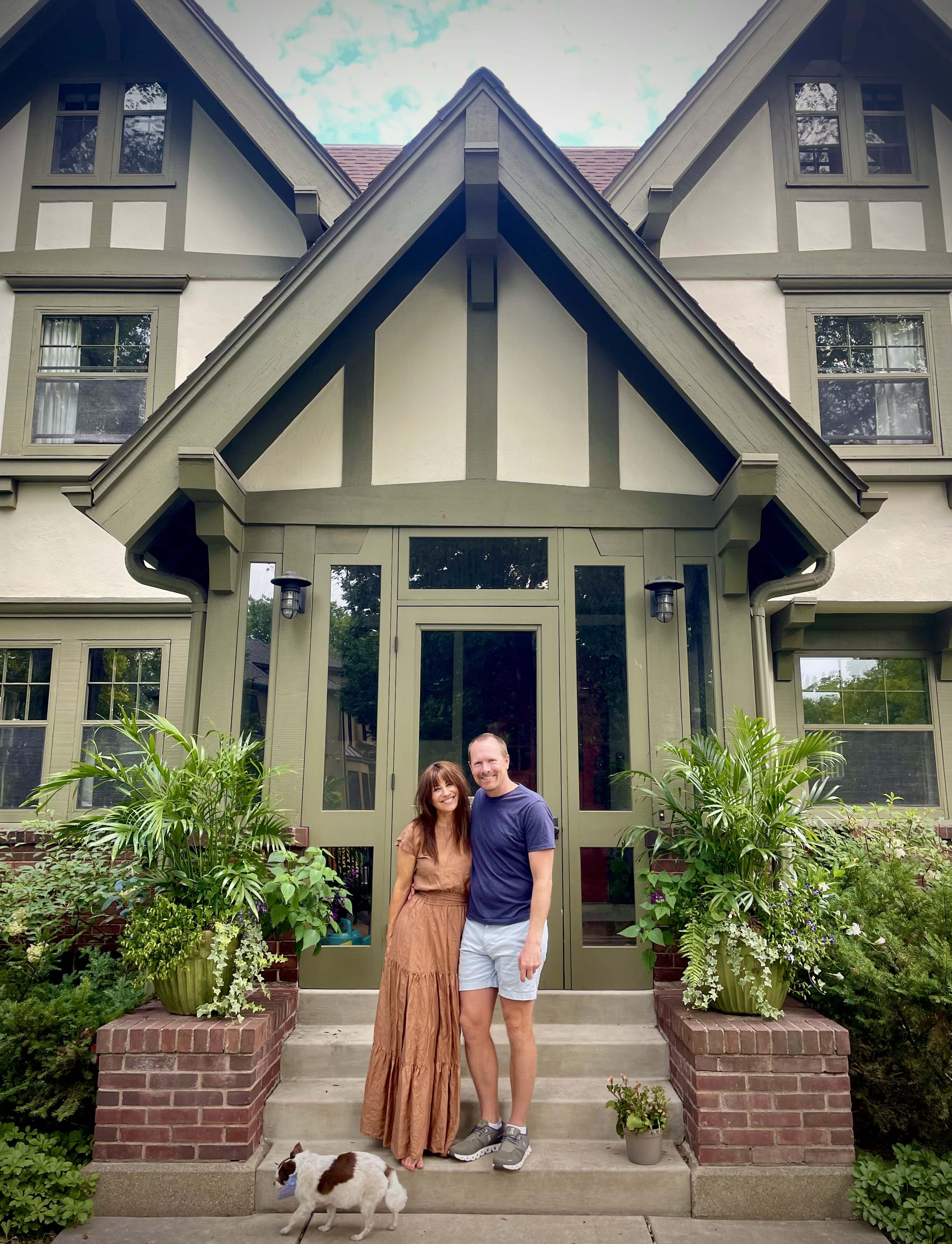 A woman in a long brown dress and a man in a gray shirt stand in front of a large, two-story house with green trim and potted plants on either side of the entrance, accompanied by a small dog.