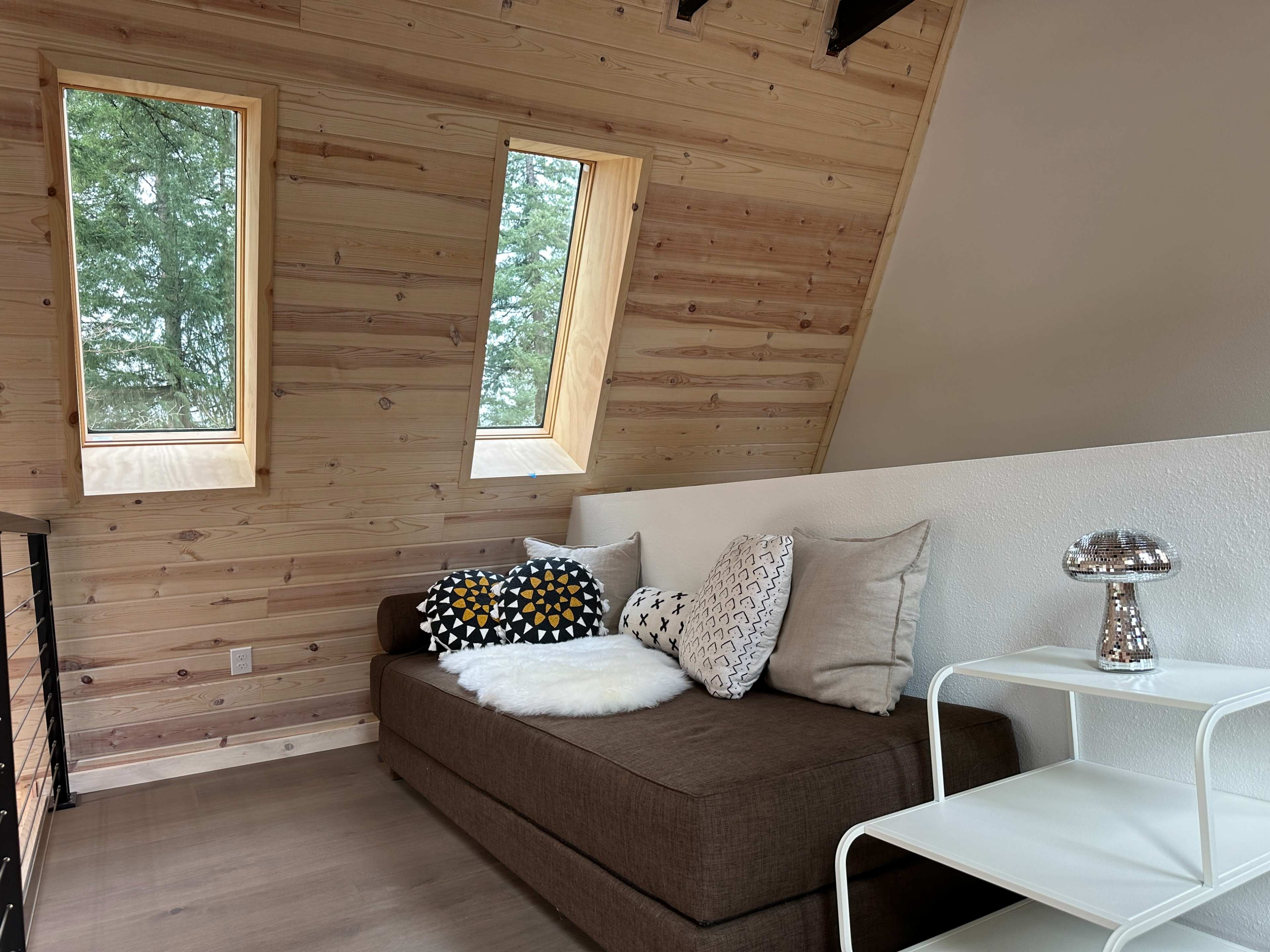 A cozy interior with a brown sectional couch adorned with decorative pillows, a small white side table, and large windows set into a wood-paneled wall.