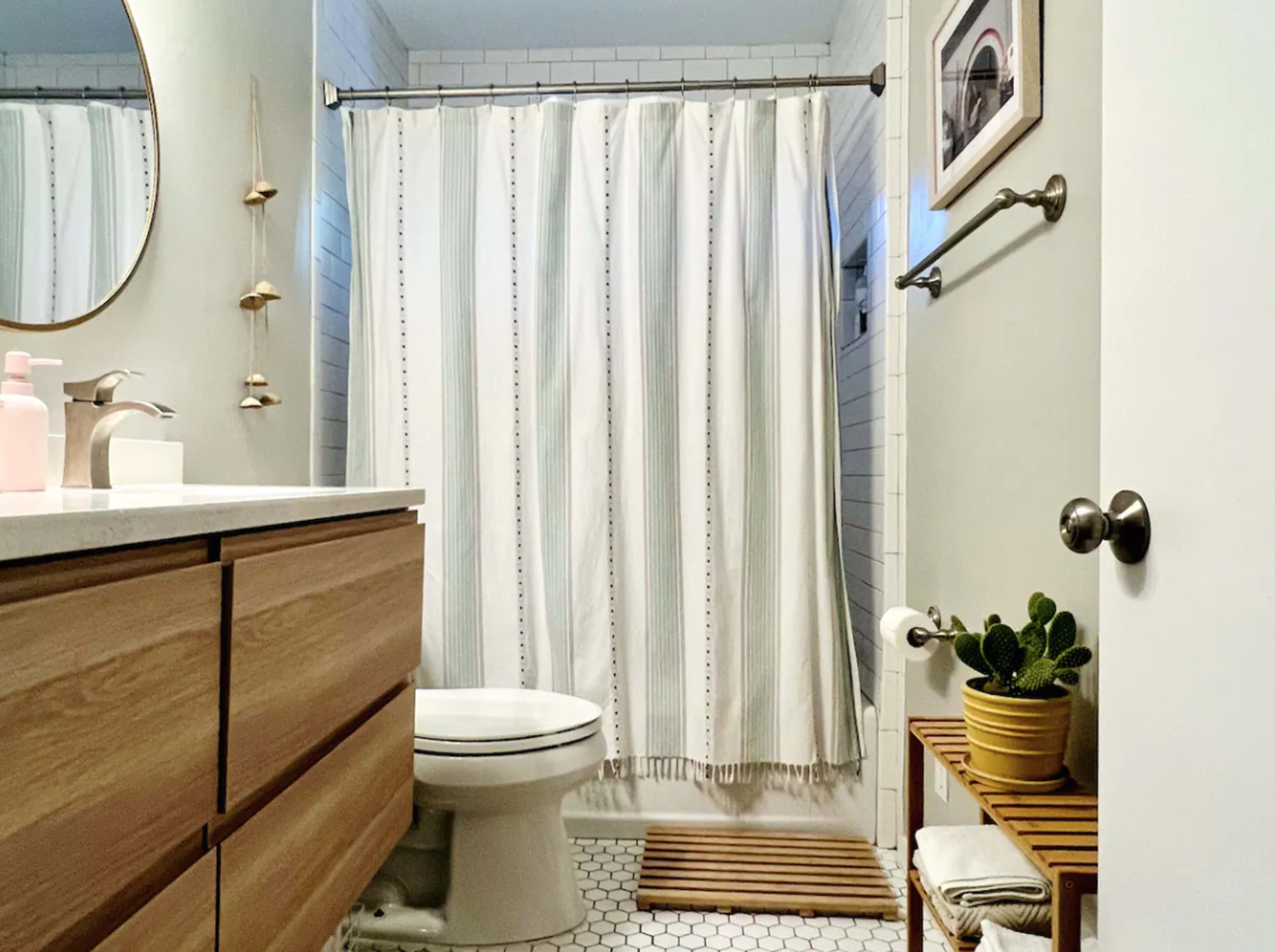 A modern bathroom features a white shower curtain, a circular mirror, a wooden vanity with storage, and potted plants on a wooden shelf.