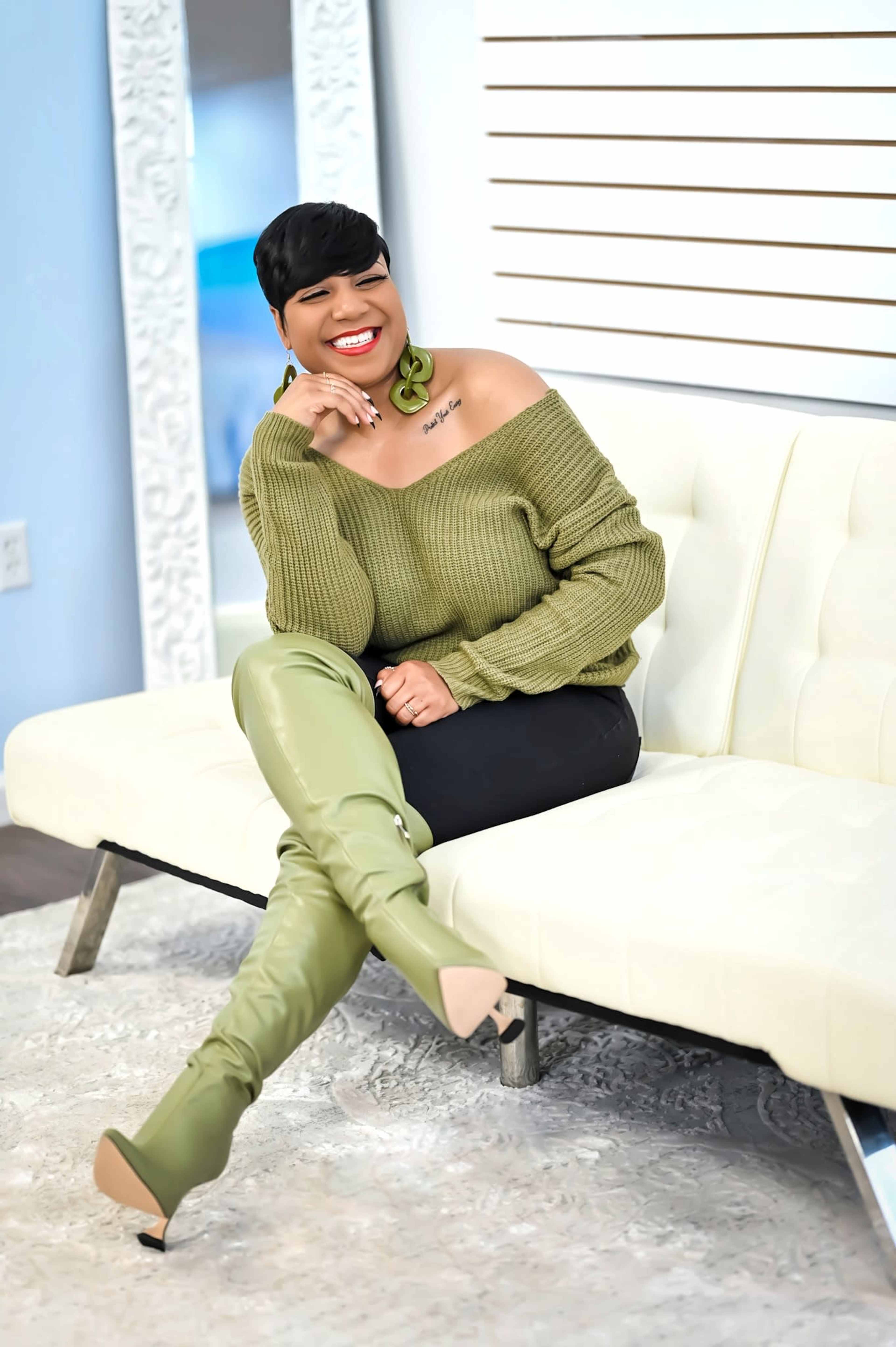 A woman wearing a green off-the-shoulder sweater and long green boots smiles while sitting on a white couch.