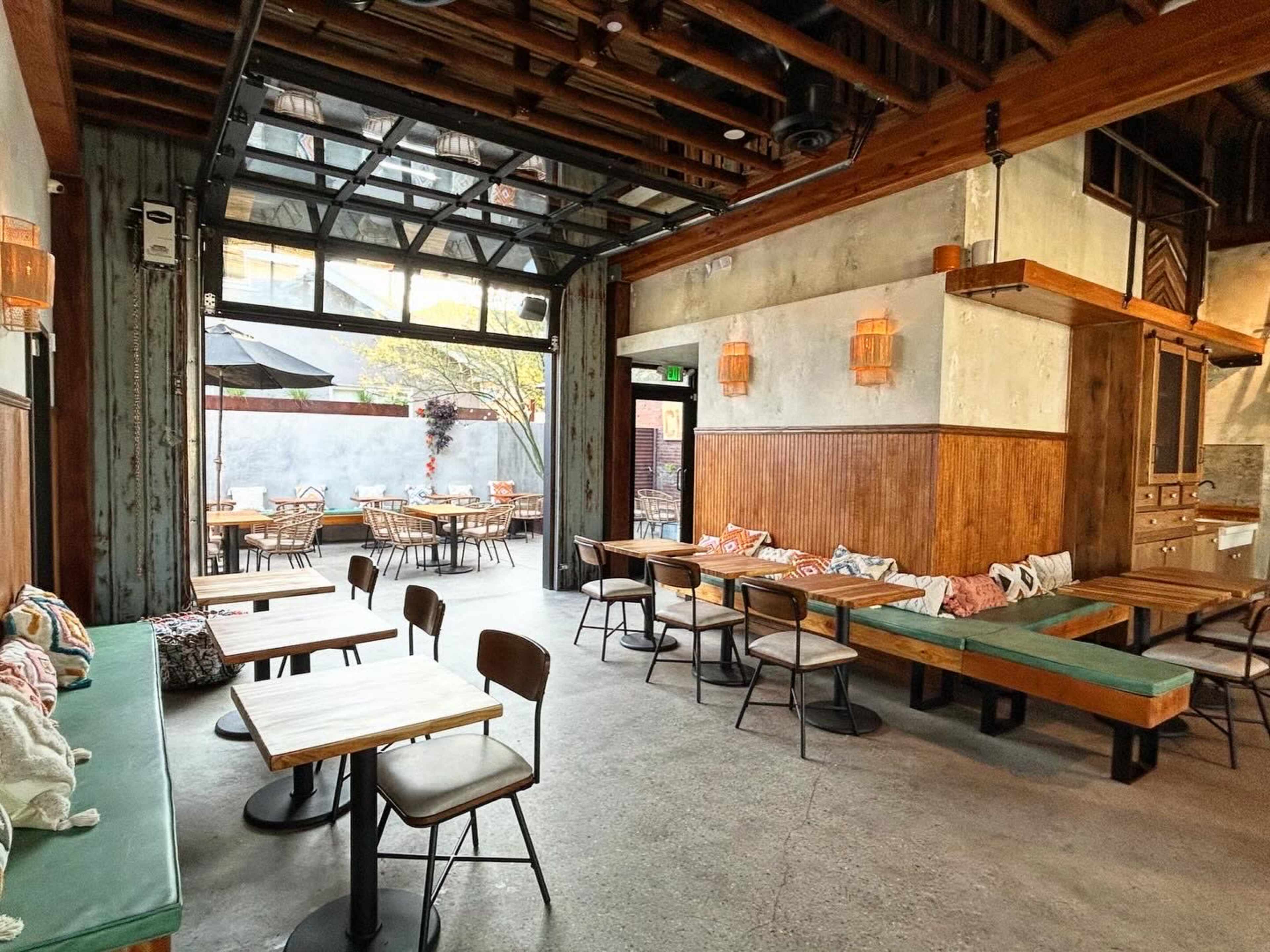The image shows a modern restaurant interior with wooden tables, benches, and large glass doors leading to an outdoor dining area.