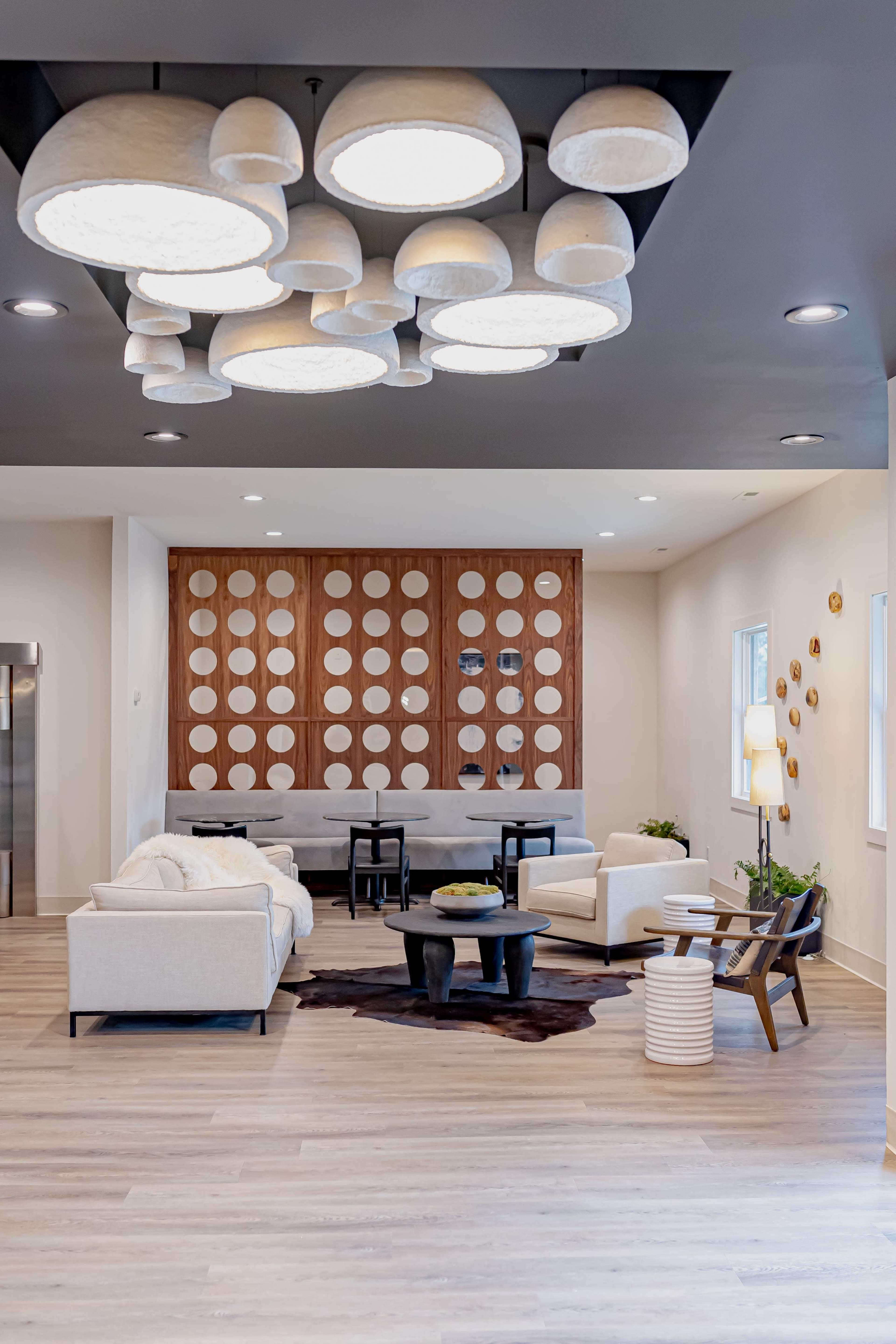 A modern lounge area with a minimalist design, featuring light-colored furniture, decorative wall panels, and a cluster of pendant lights overhead.