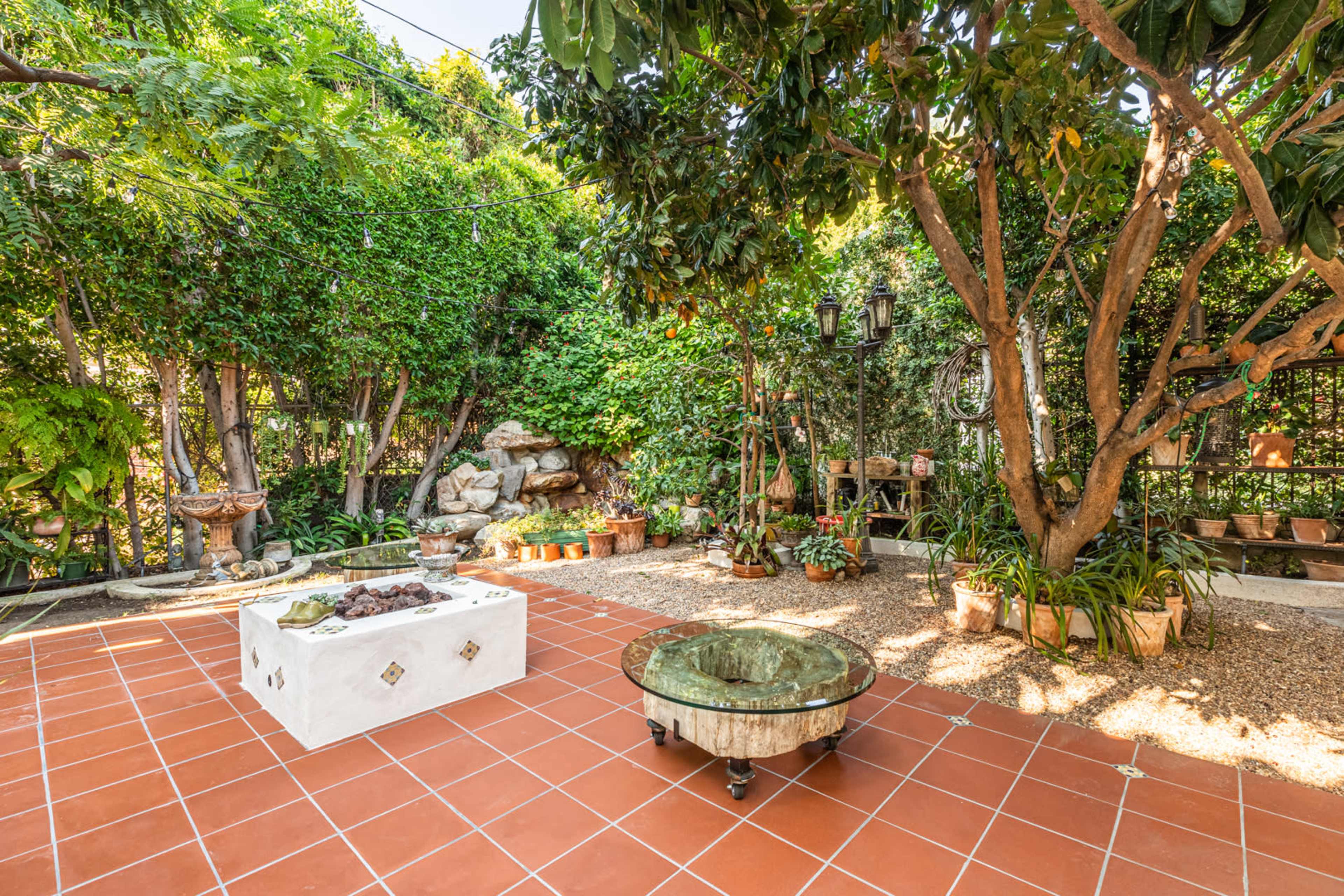 A garden area with a tiled patio featuring a stone table and a glass-top coffee table surrounded by lush green plants and trees.