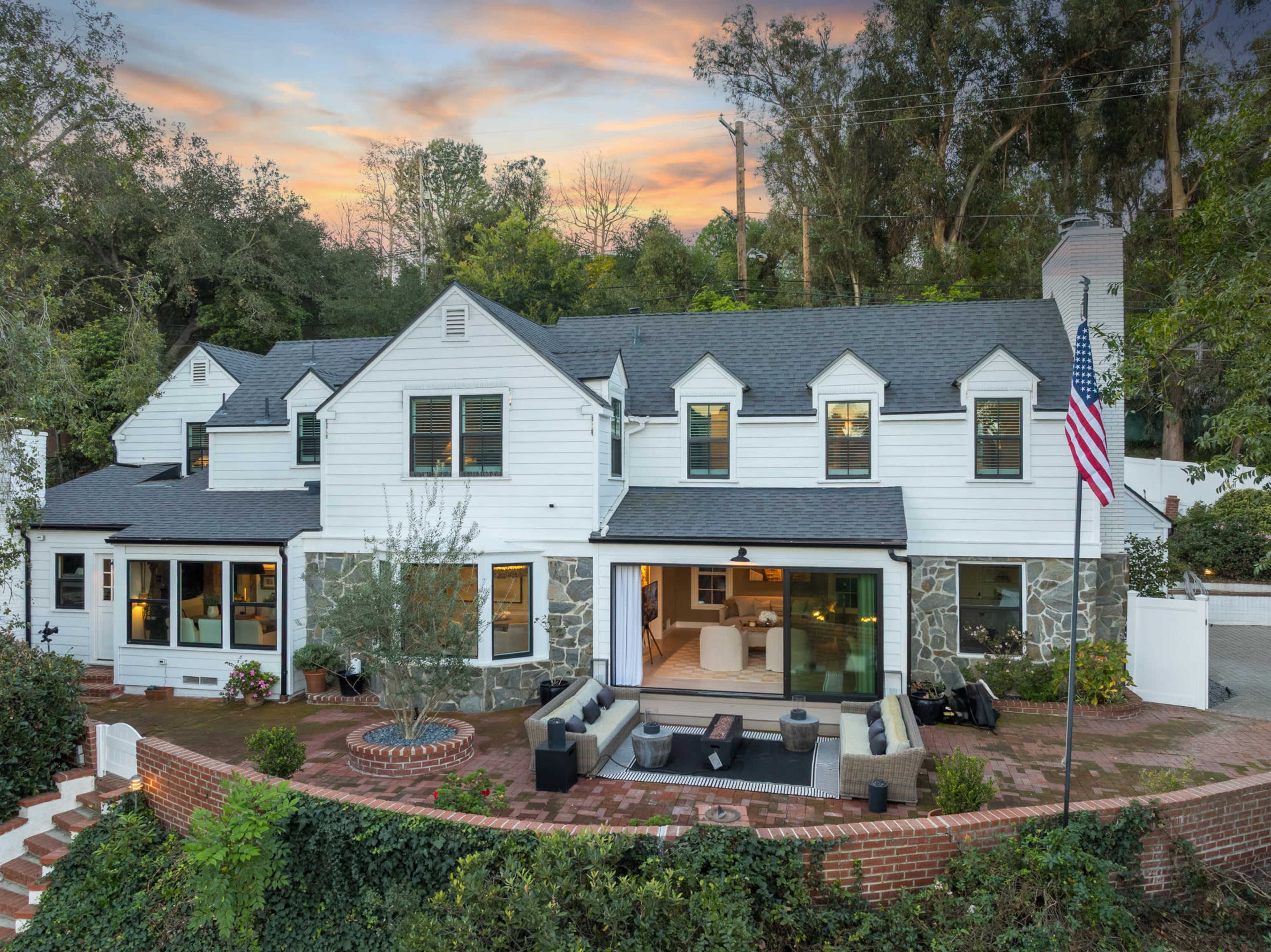A two-story house with a white exterior and stone accents features a spacious outdoor seating area and is surrounded by trees, set against a vibrant sunset sky.