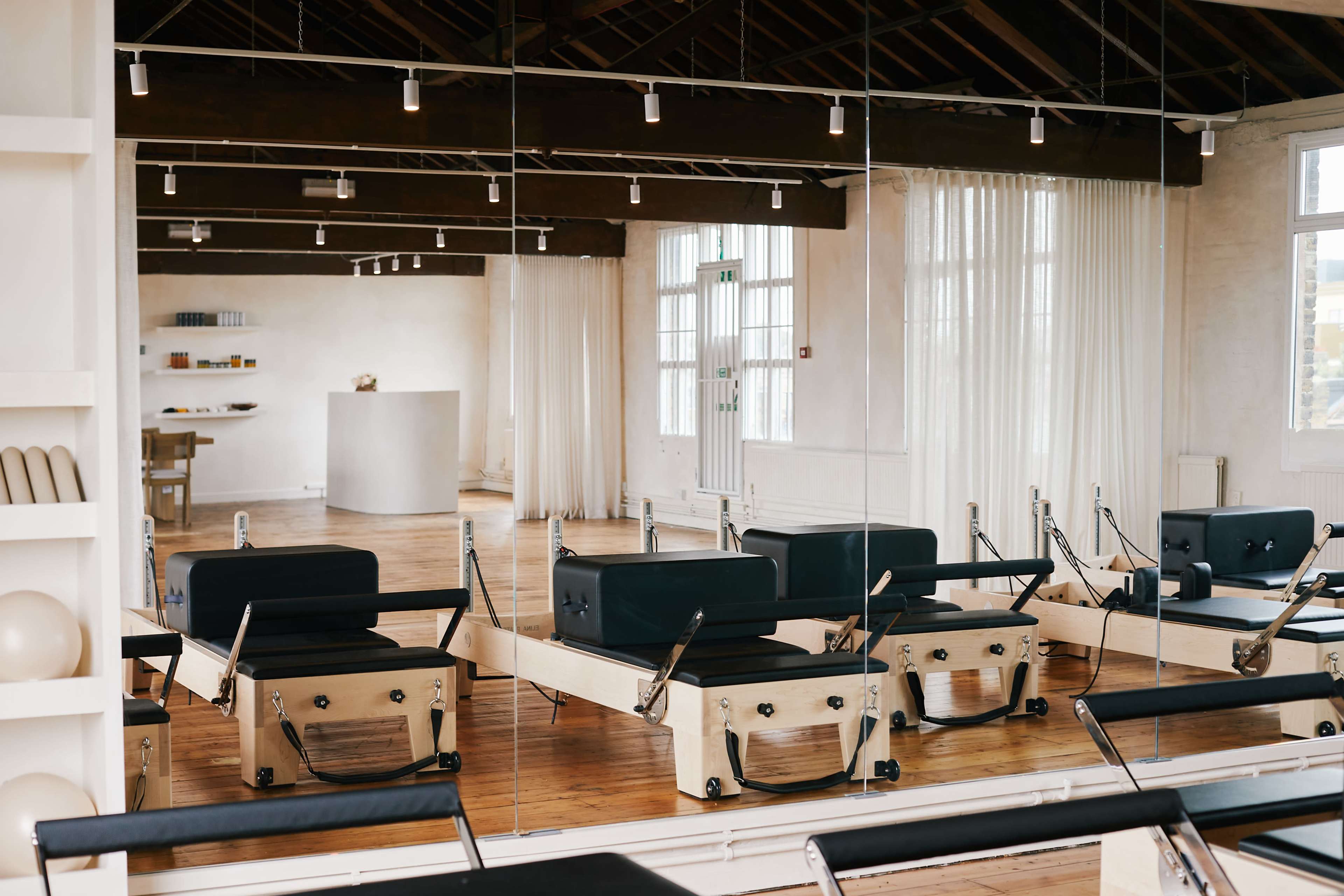 The image shows a spacious Pilates studio with reformers arranged in front of large mirrors and natural light streaming in through the windows.