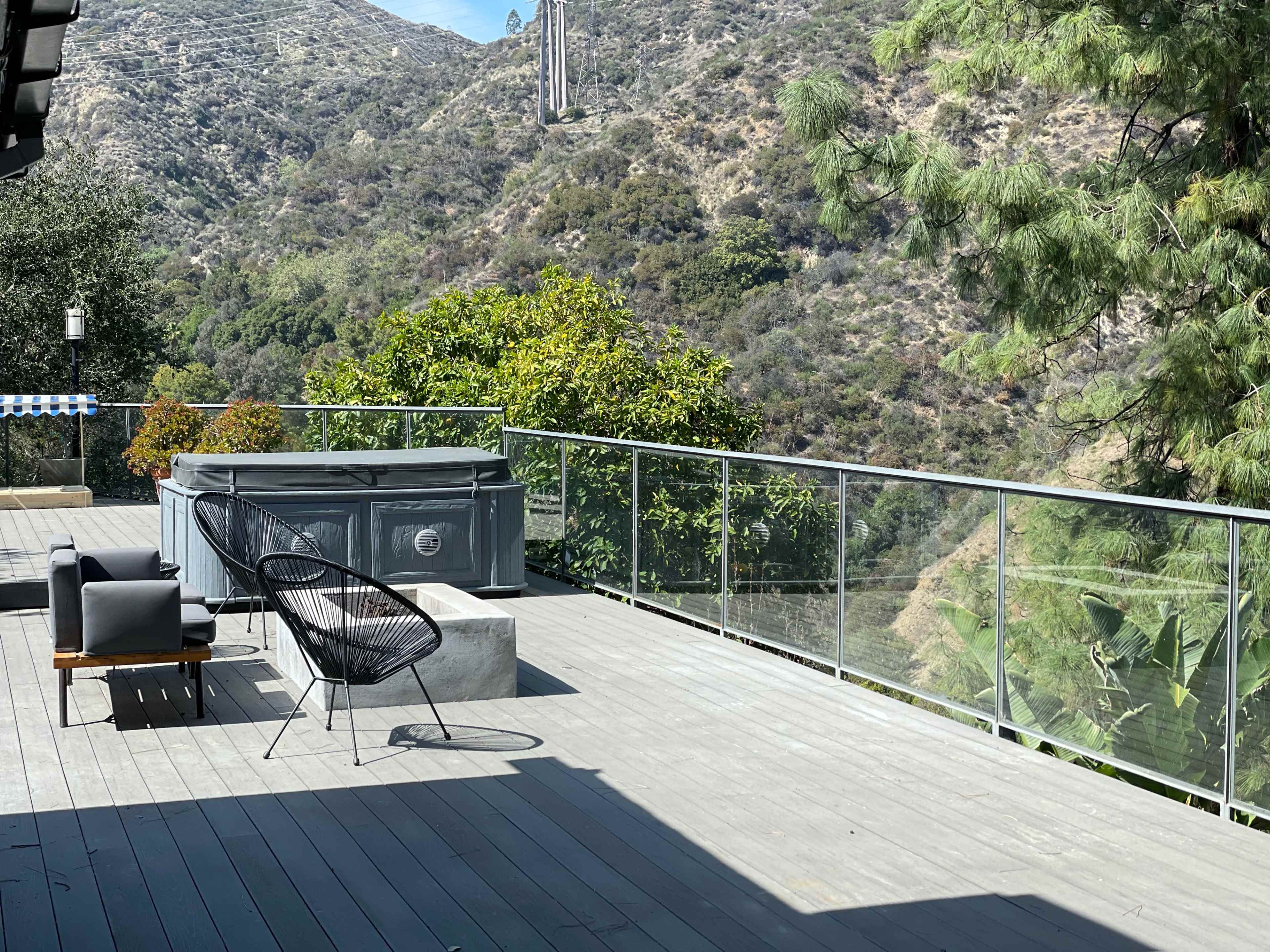 A deck overlooks a mountainous landscape, featuring a hot tub, an outdoor seating area, and greenery in the background.