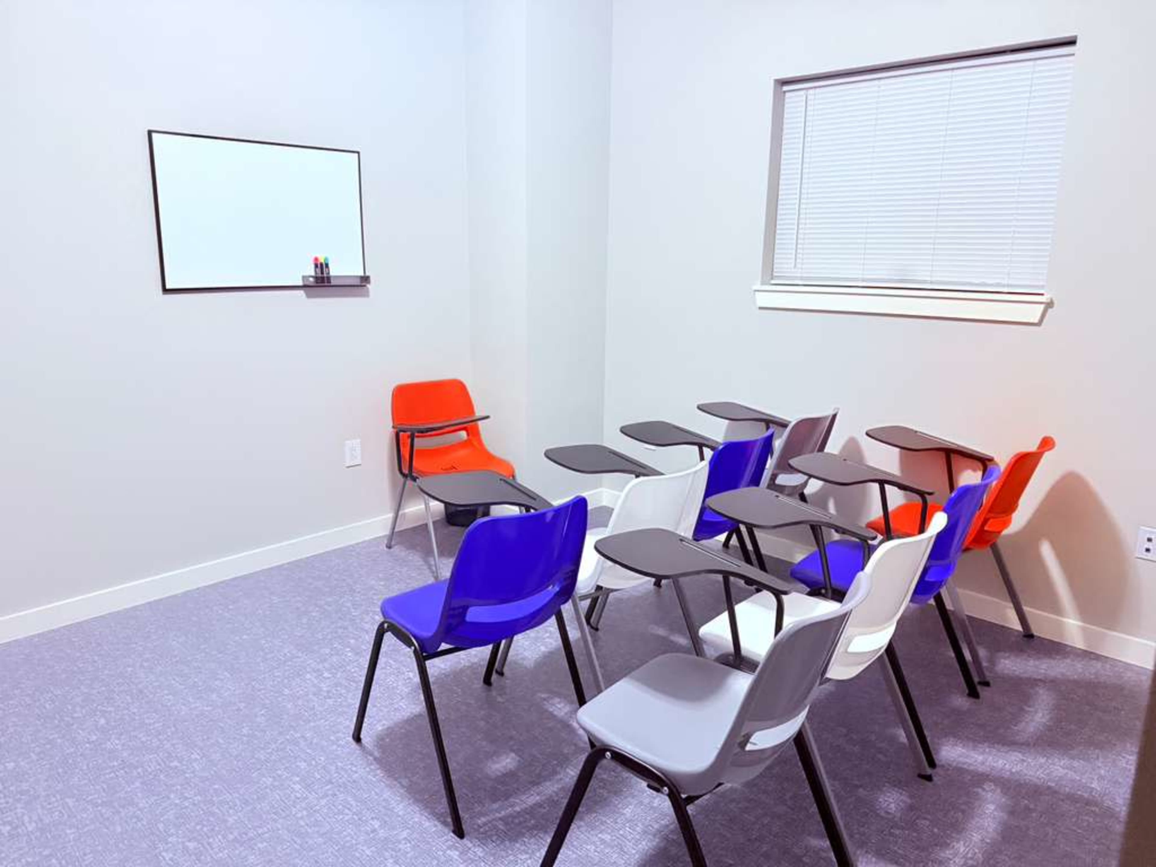 A small classroom is arranged with several chairs and desks in various colors, facing a wall with a whiteboard and markers.
