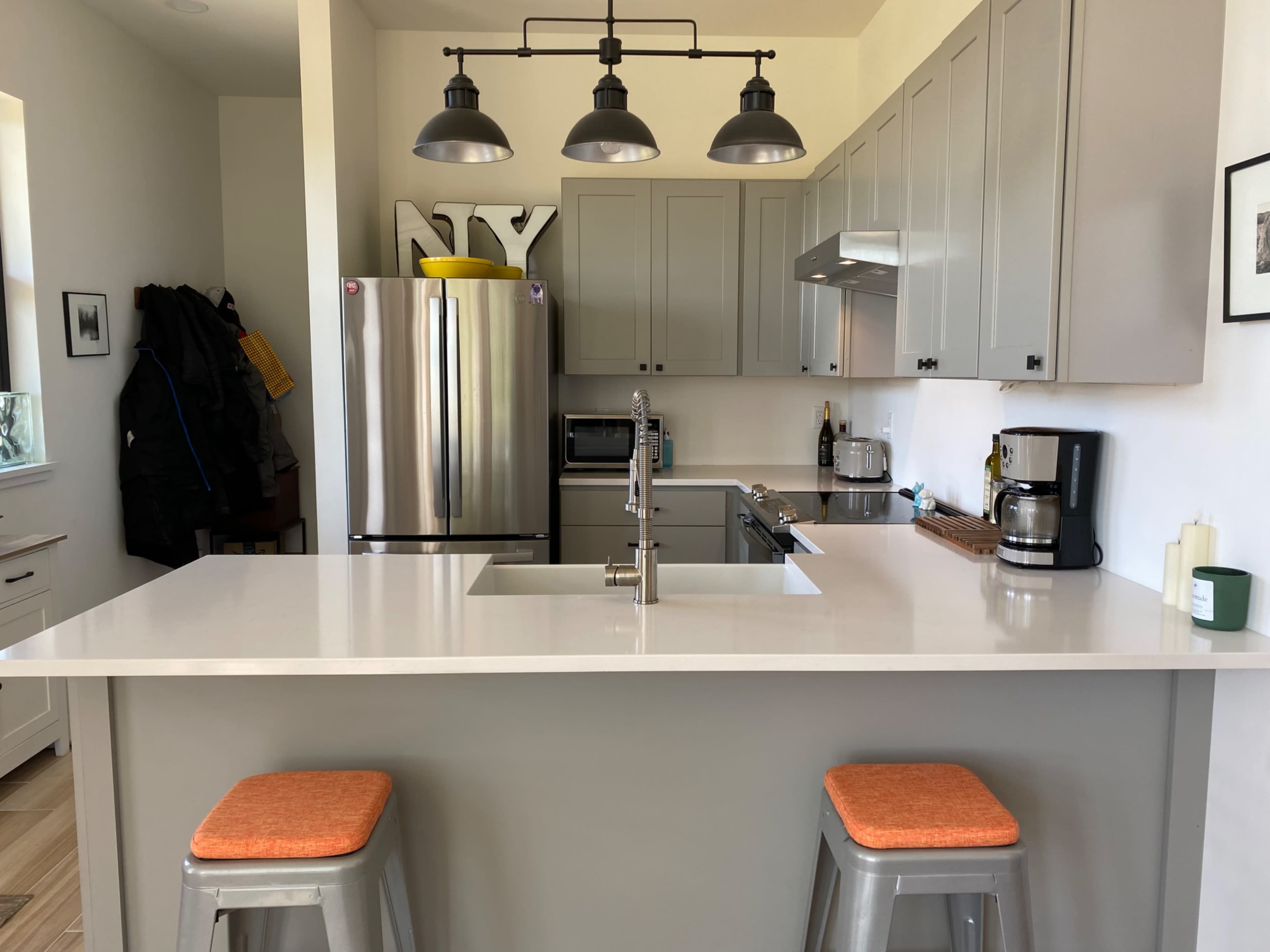 A modern kitchen with gray cabinets, stainless steel appliances, a large island with a white countertop, and two orange stools.