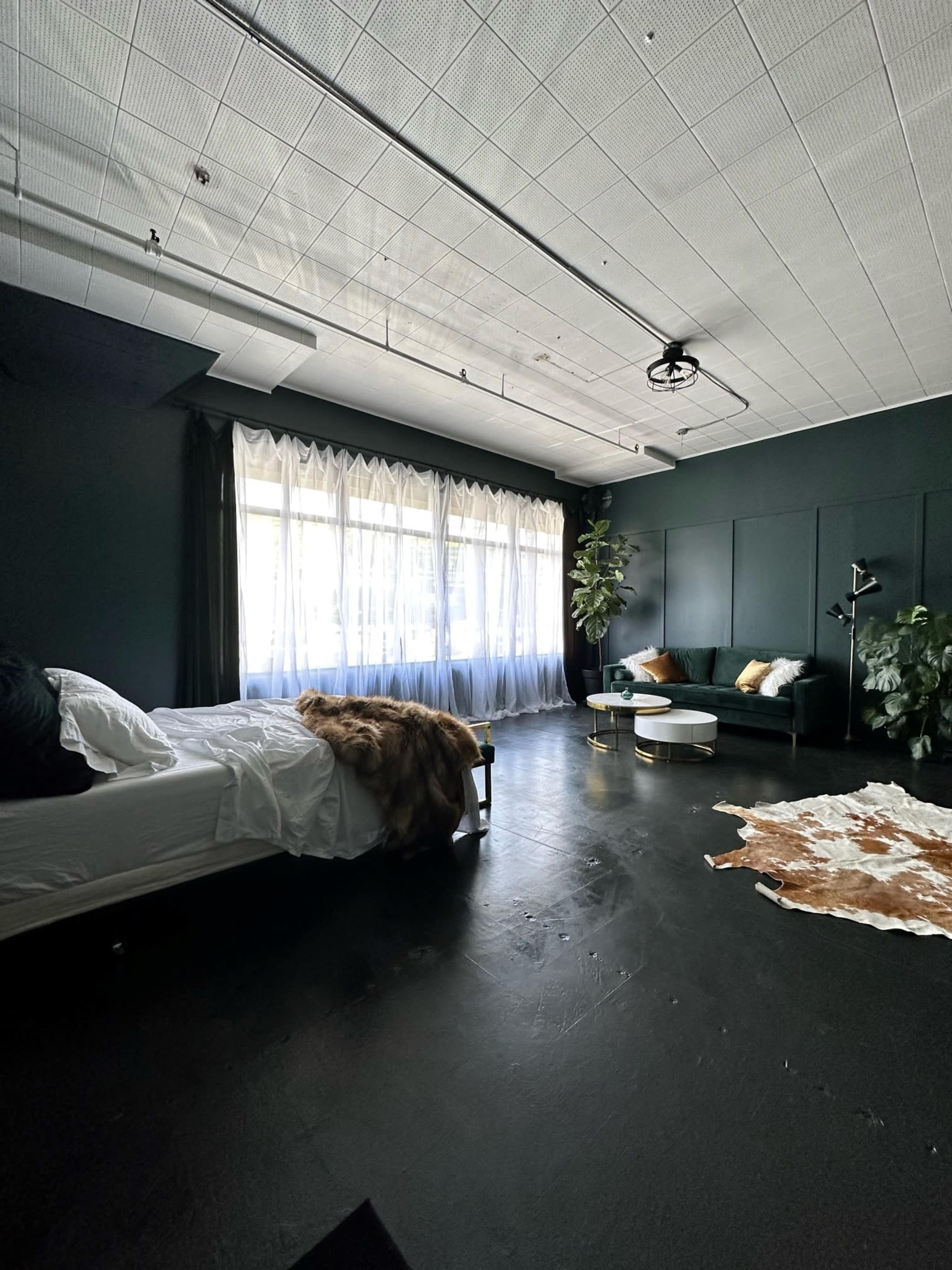 The image shows a dark-themed bedroom with a bed, a sofa, a large window covered with sheer curtains, and a decorative rug on the floor.