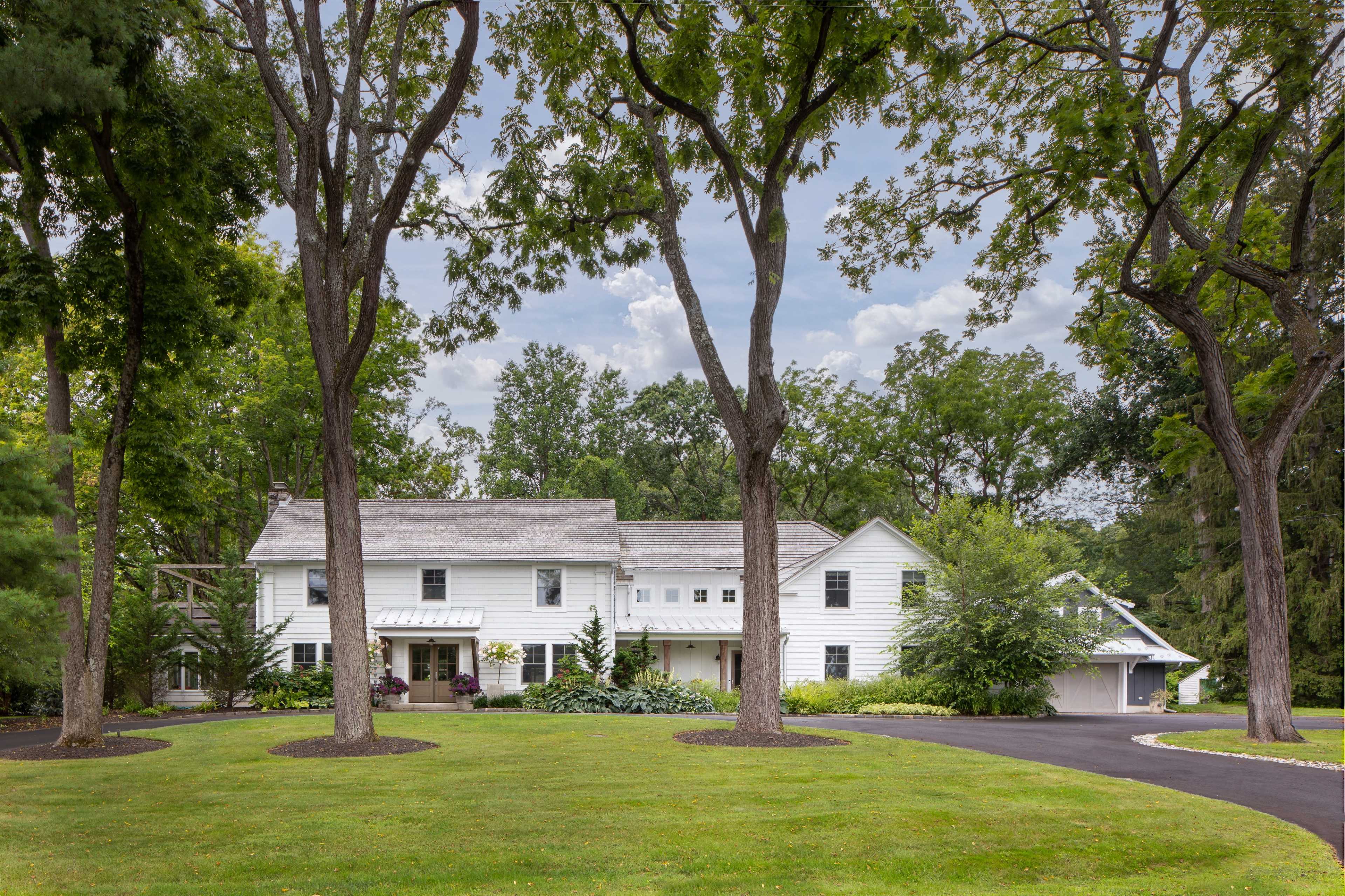 A large, white two-story house is situated among tall trees on a manicured lawn, with a paved driveway leading up to it.