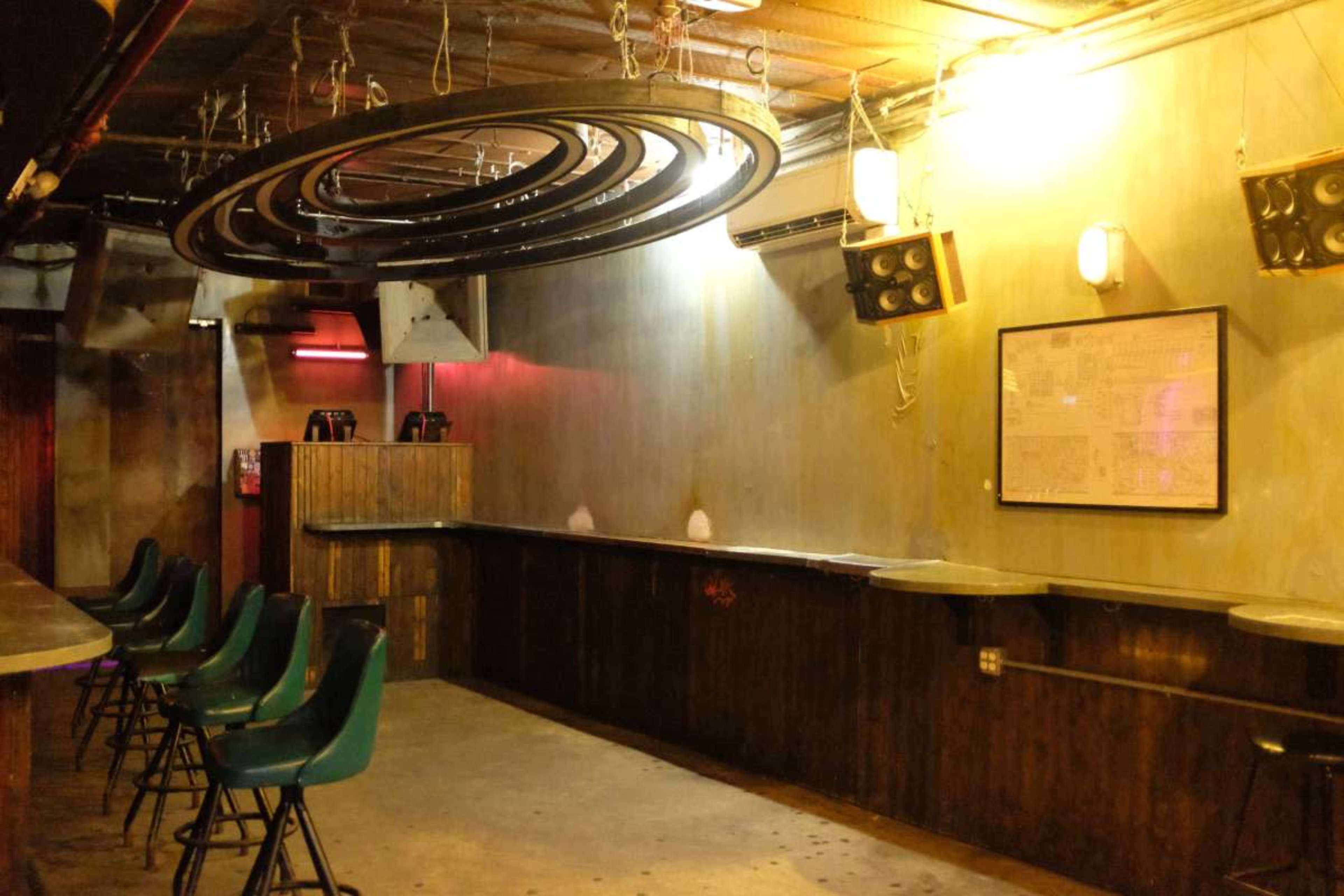 The image shows an empty bar area with a long wooden counter, green stools, and circular light fixtures hanging from the ceiling.