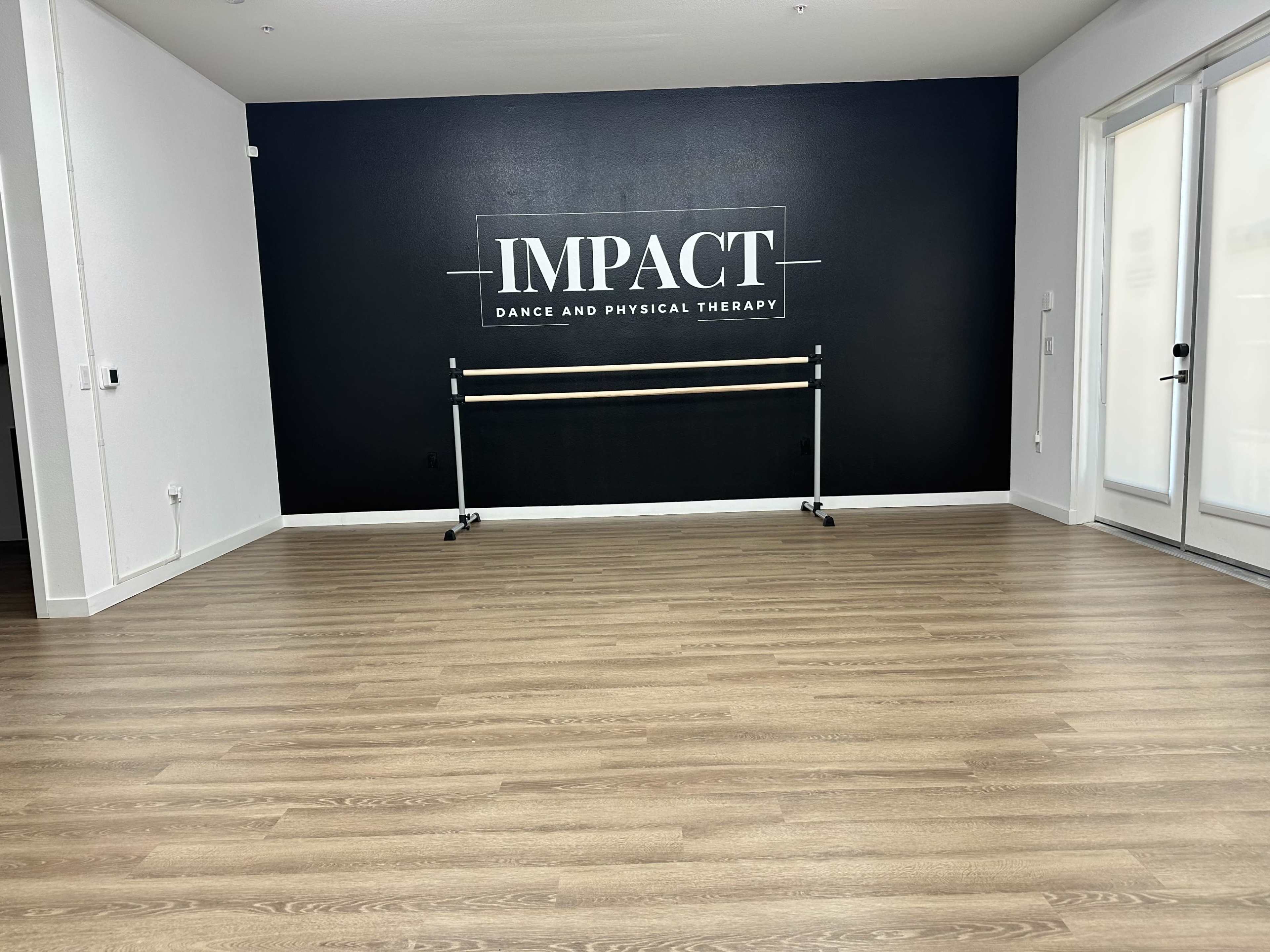 A studio space with light wooden flooring and a black wall featuring the words "IMPACT DANCE AND PHYSICAL THERAPY," along with a ballet barre on wheels.