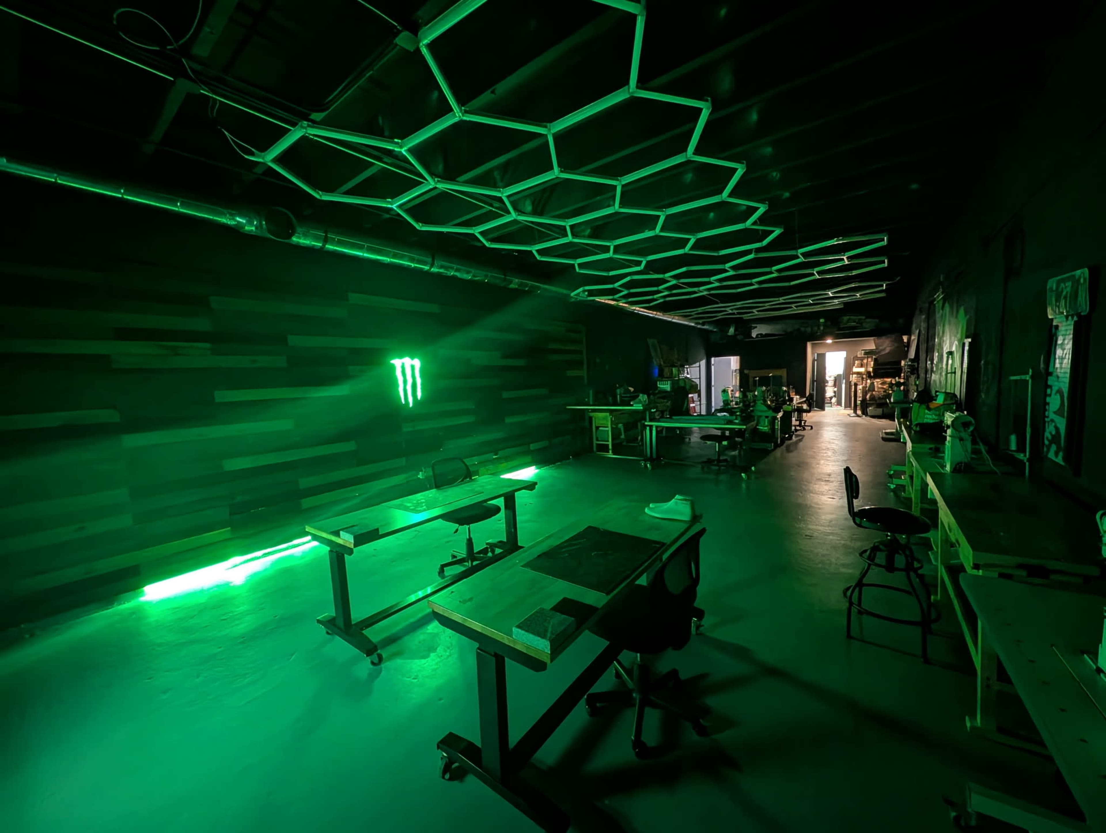 The image shows a spacious workspace with a dark wall, green LED lighting, and several tables and chairs arranged throughout.