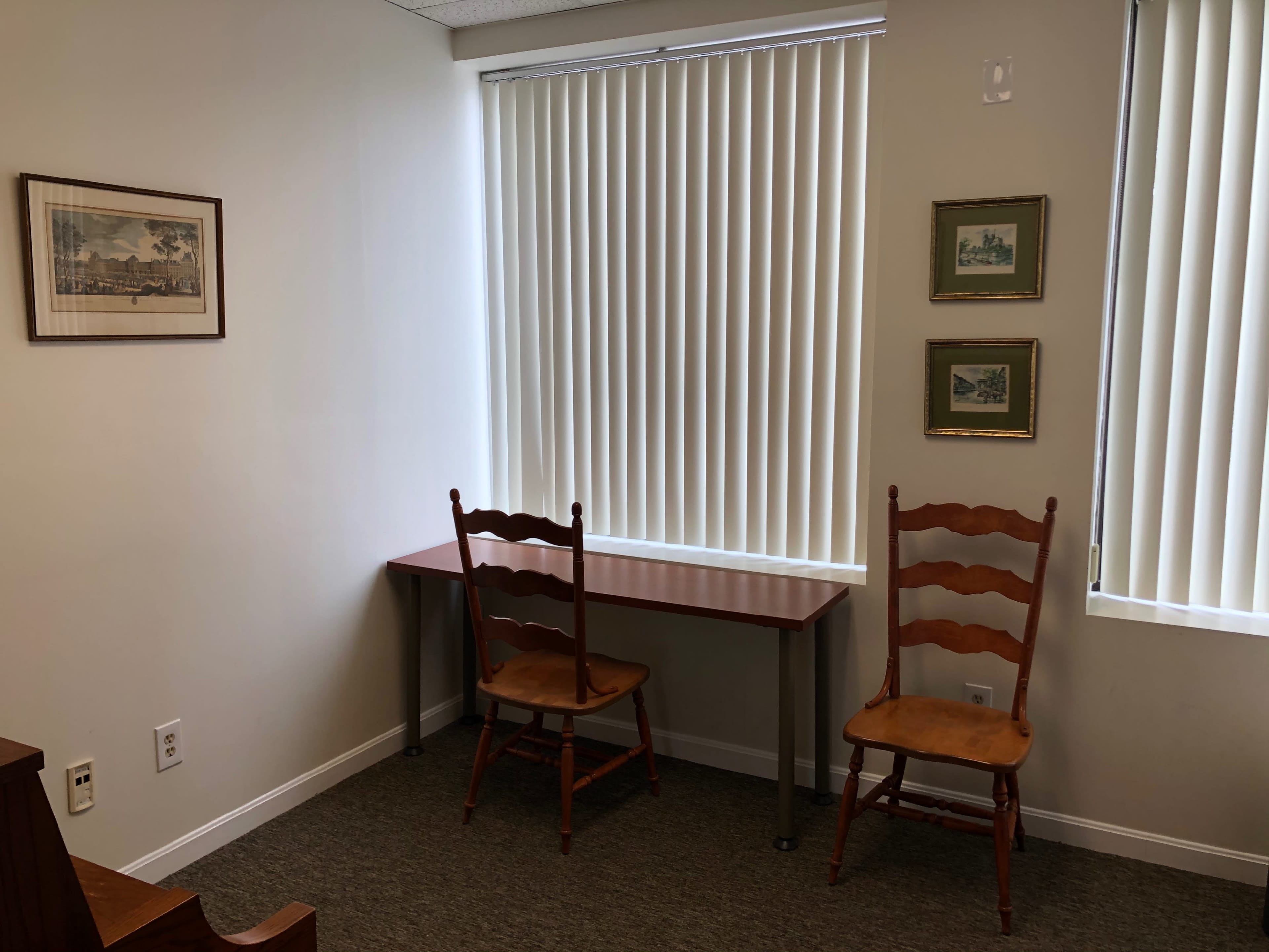 A small office space with a table and two wooden chairs, positioned near windows with vertical blinds, and framed artwork on the walls.