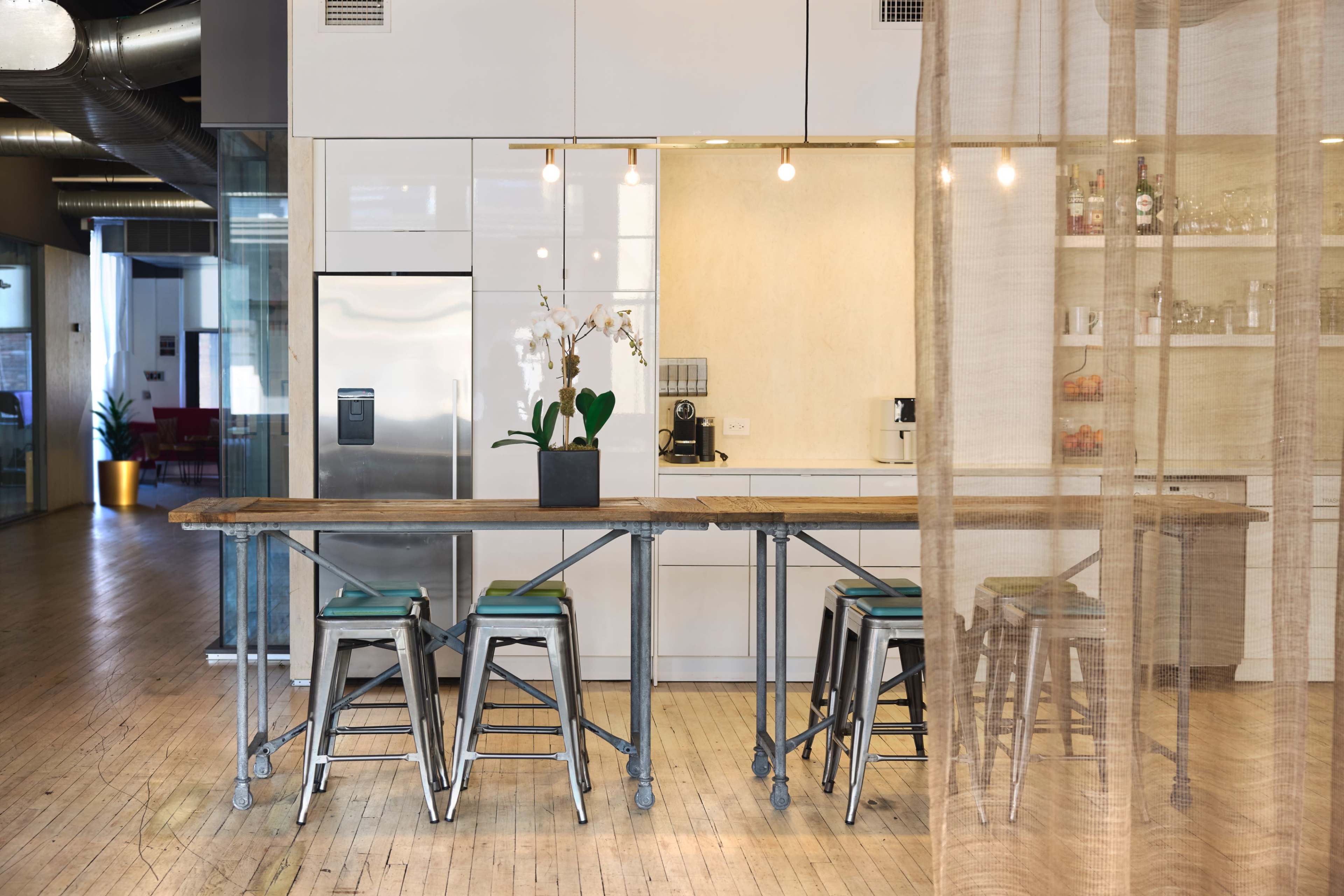 A modern kitchen area features a long wooden table with metal stools, surrounded by white cabinetry and a glimpse of a bar with beverages in the background.