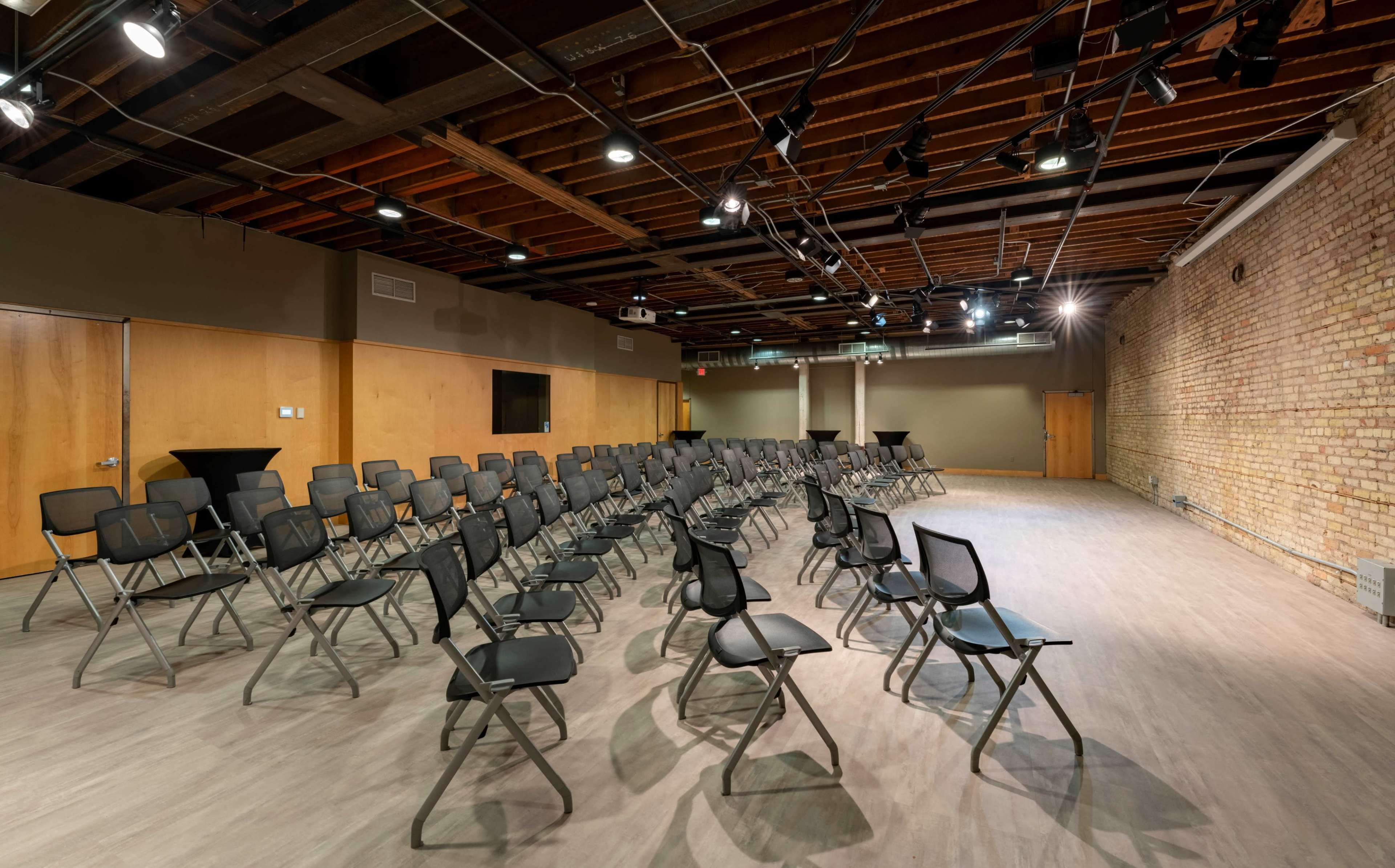 A row of black folding chairs is arranged in a spacious, well-lit auditorium with exposed wooden beams and a brick wall.