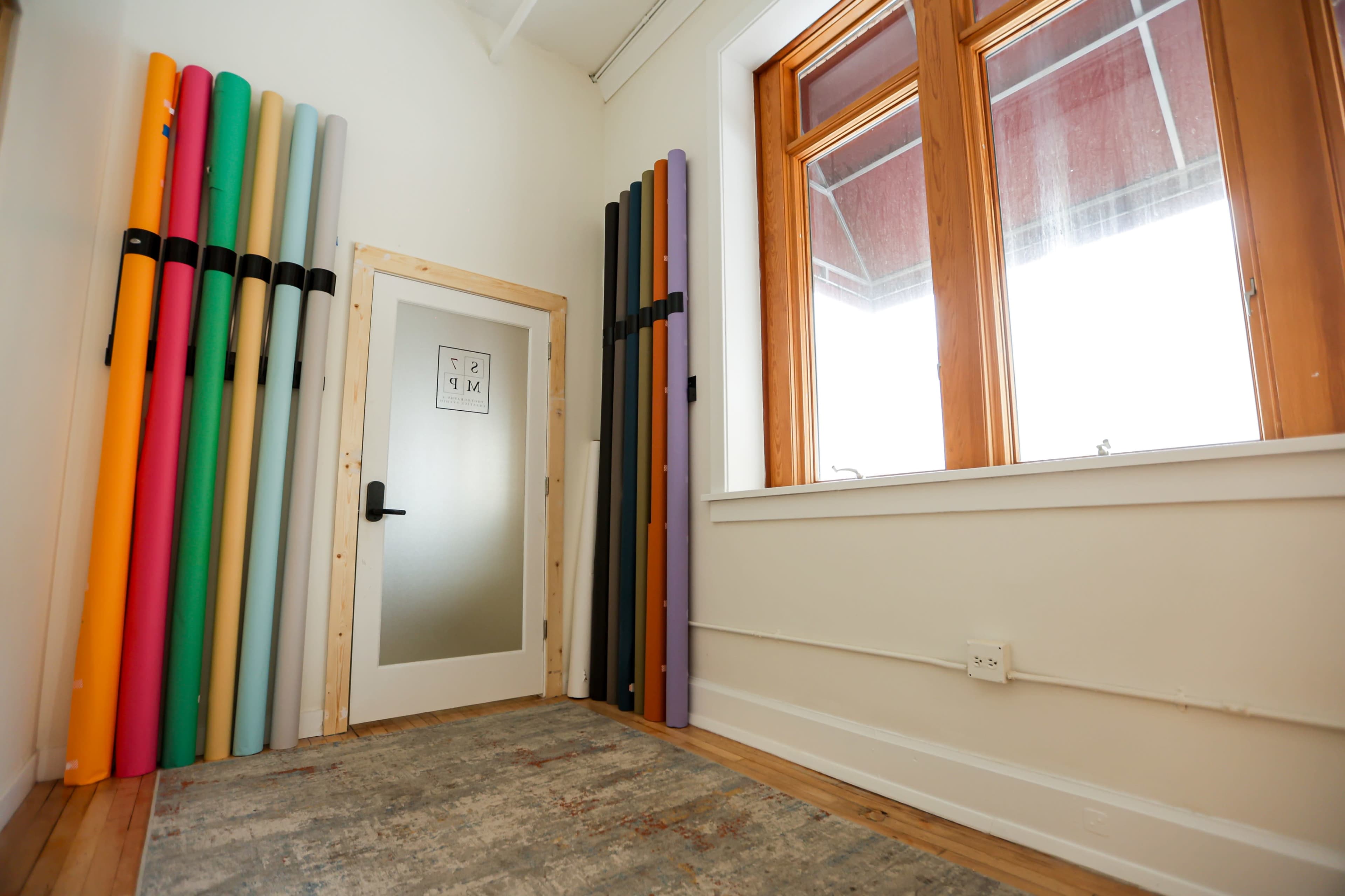The image shows a bright room with a door and a wall adorned with several rolled yoga mats neatly arranged in various colors.