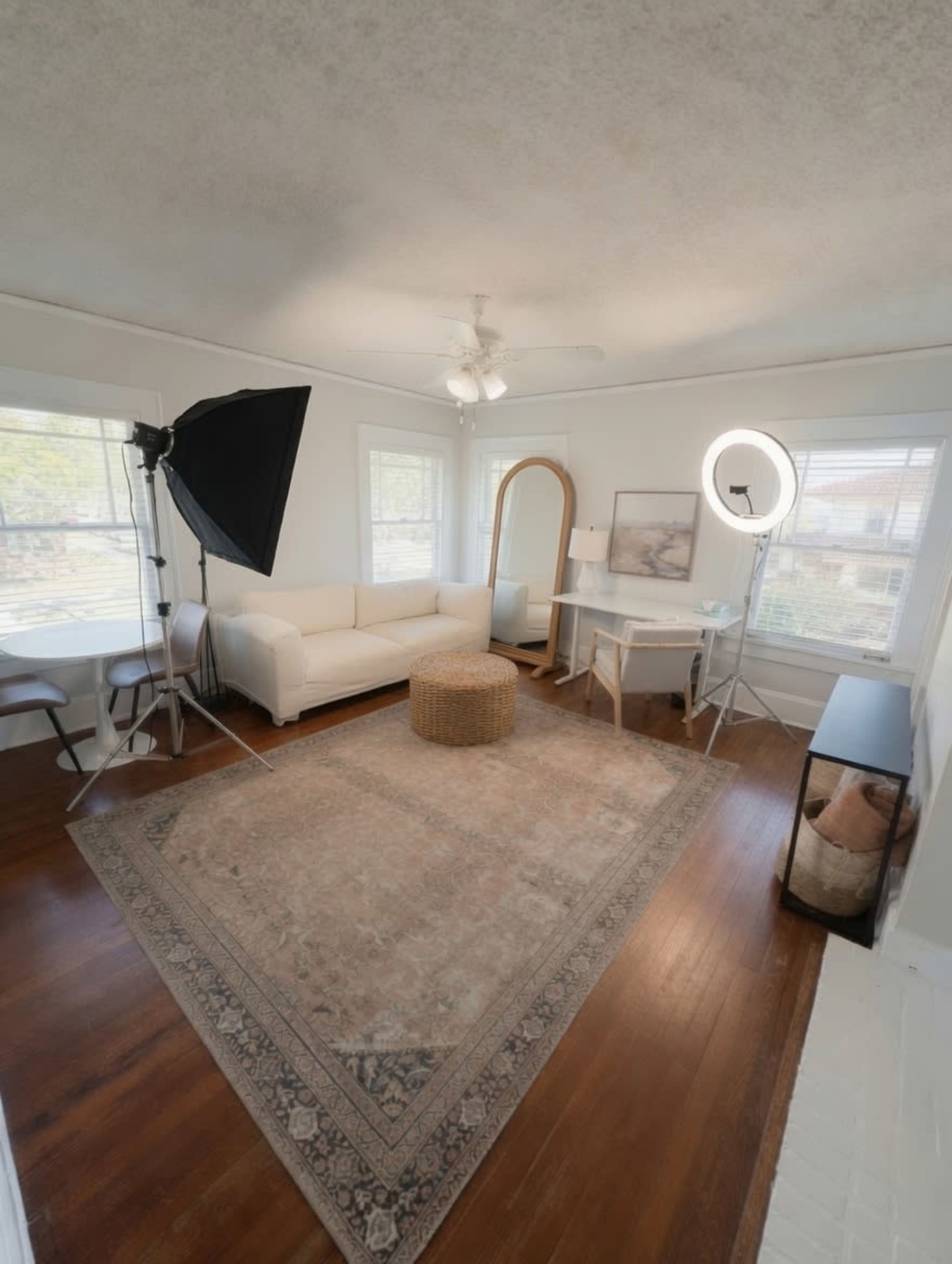 The image shows a bright, airy living room featuring a white couch, a round ottoman, a mirror, and a photography setup with a softbox and a ring light.
