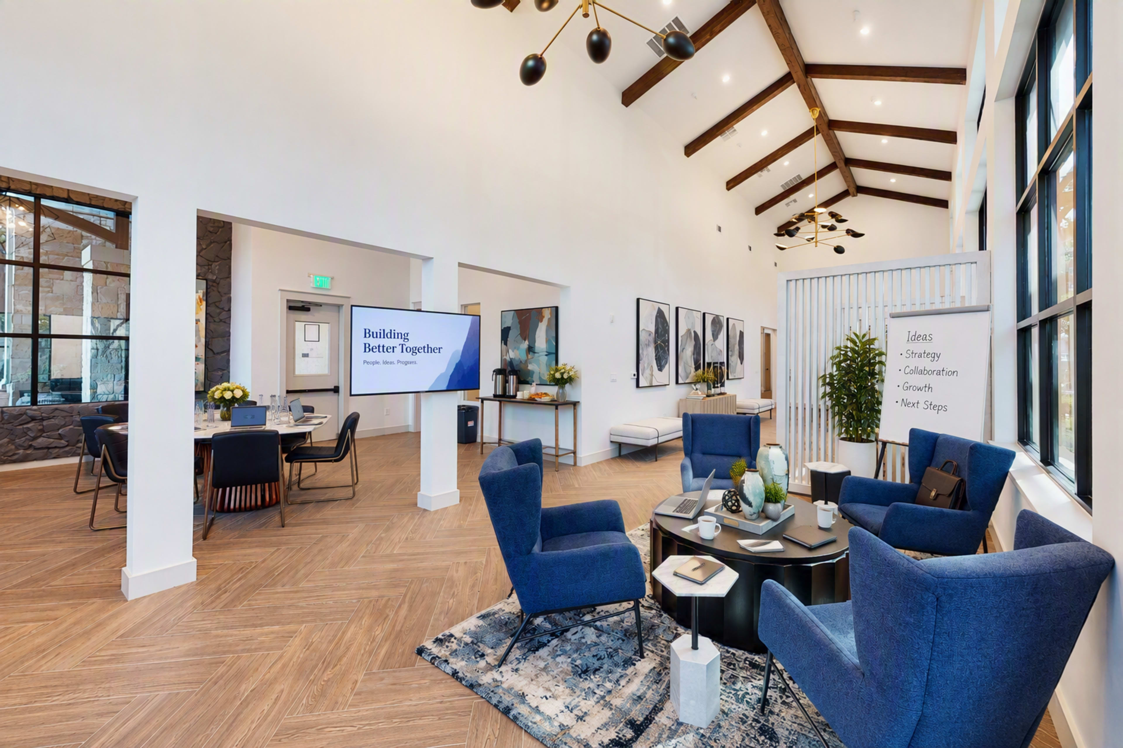 The image shows a spacious lobby with high ceilings, featuring blue armchairs, a round dining table, and decorative plants, along with a large presentation screen displaying a message about collaboration.