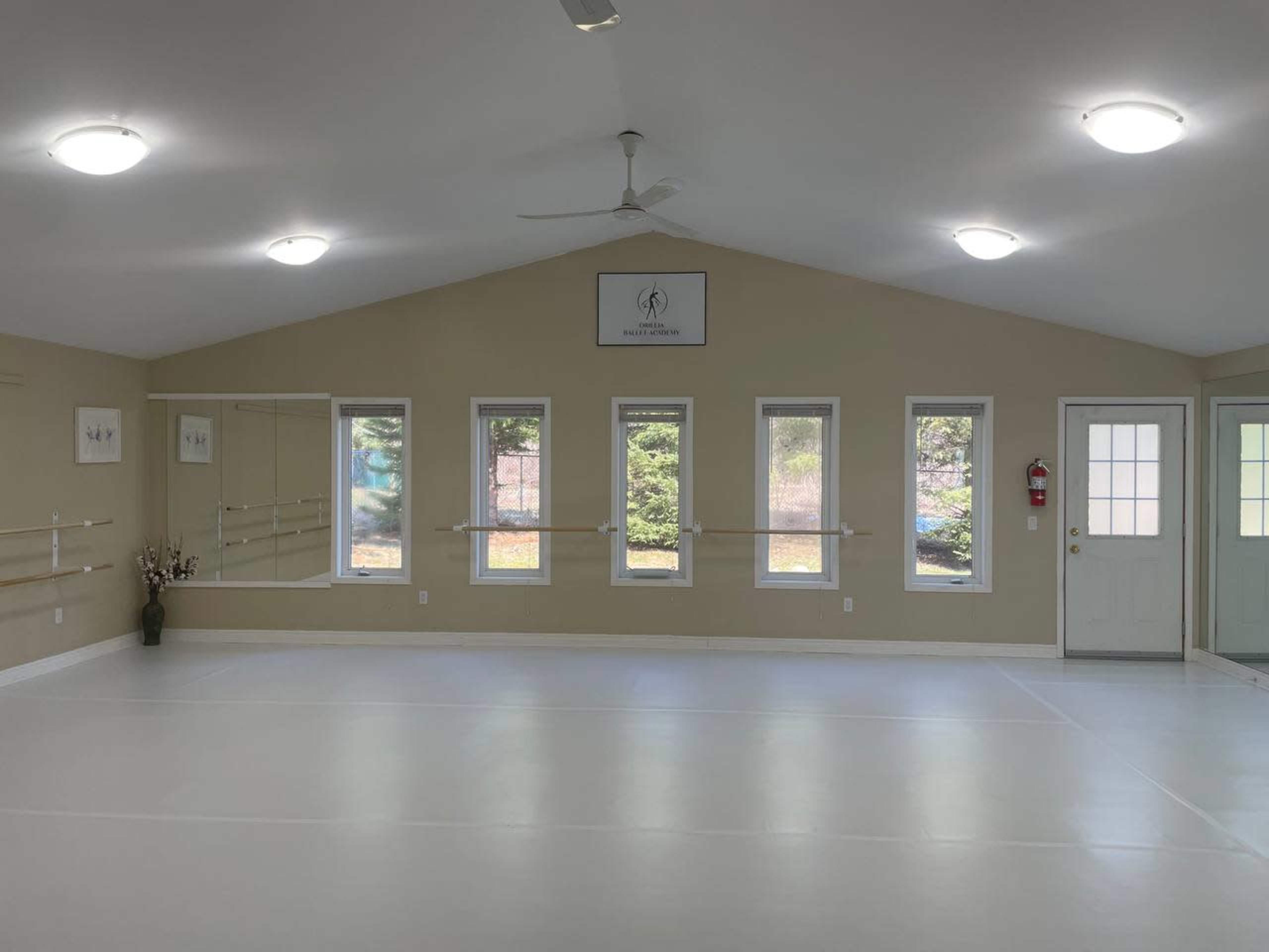 An empty dance studio with large windows, a mirror on one wall, and wooden ballet bars along another wall, illuminated by overhead lights.