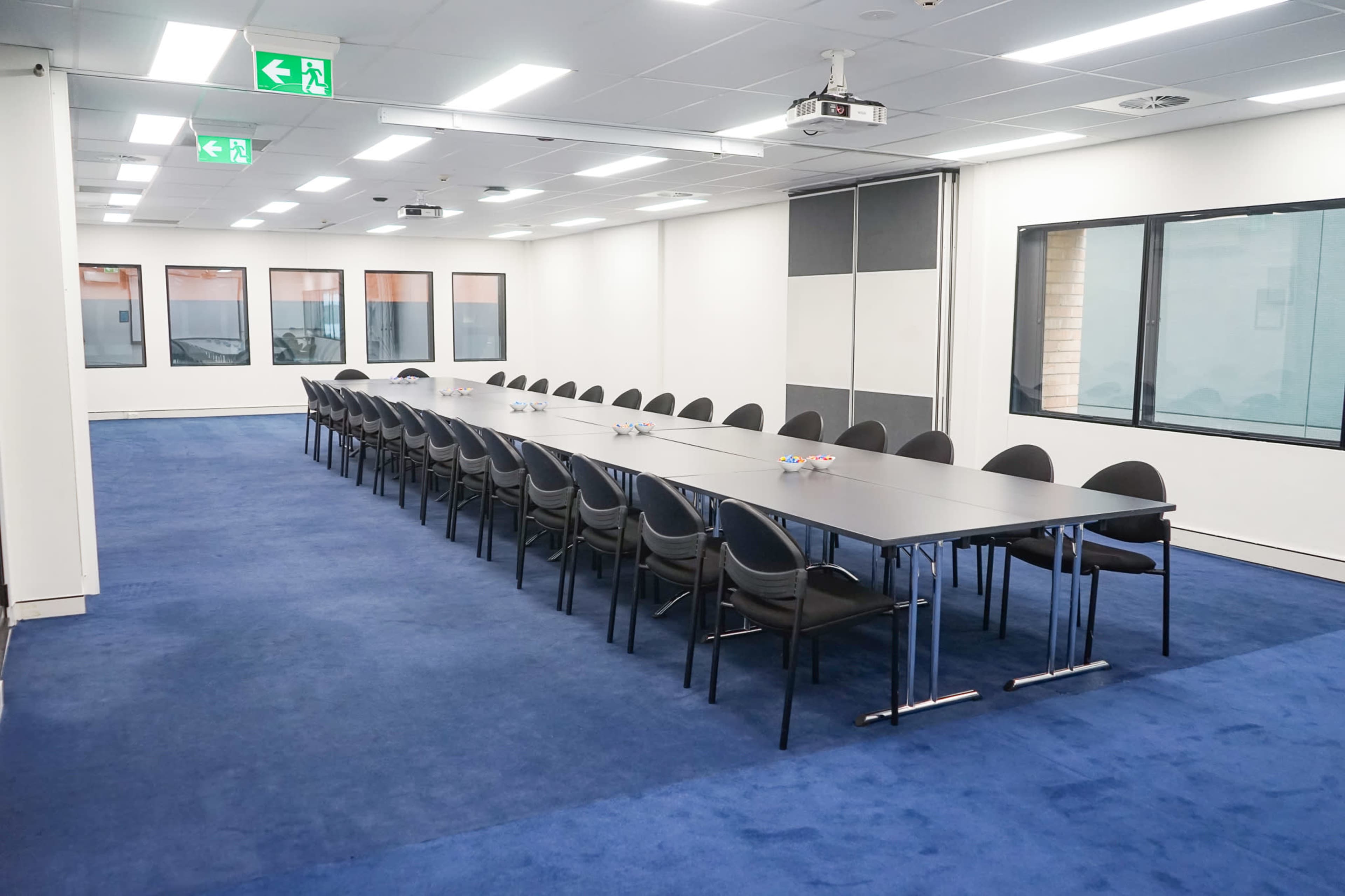 A long conference table with black chairs is arranged in a well-lit meeting room featuring windows and blue carpet.