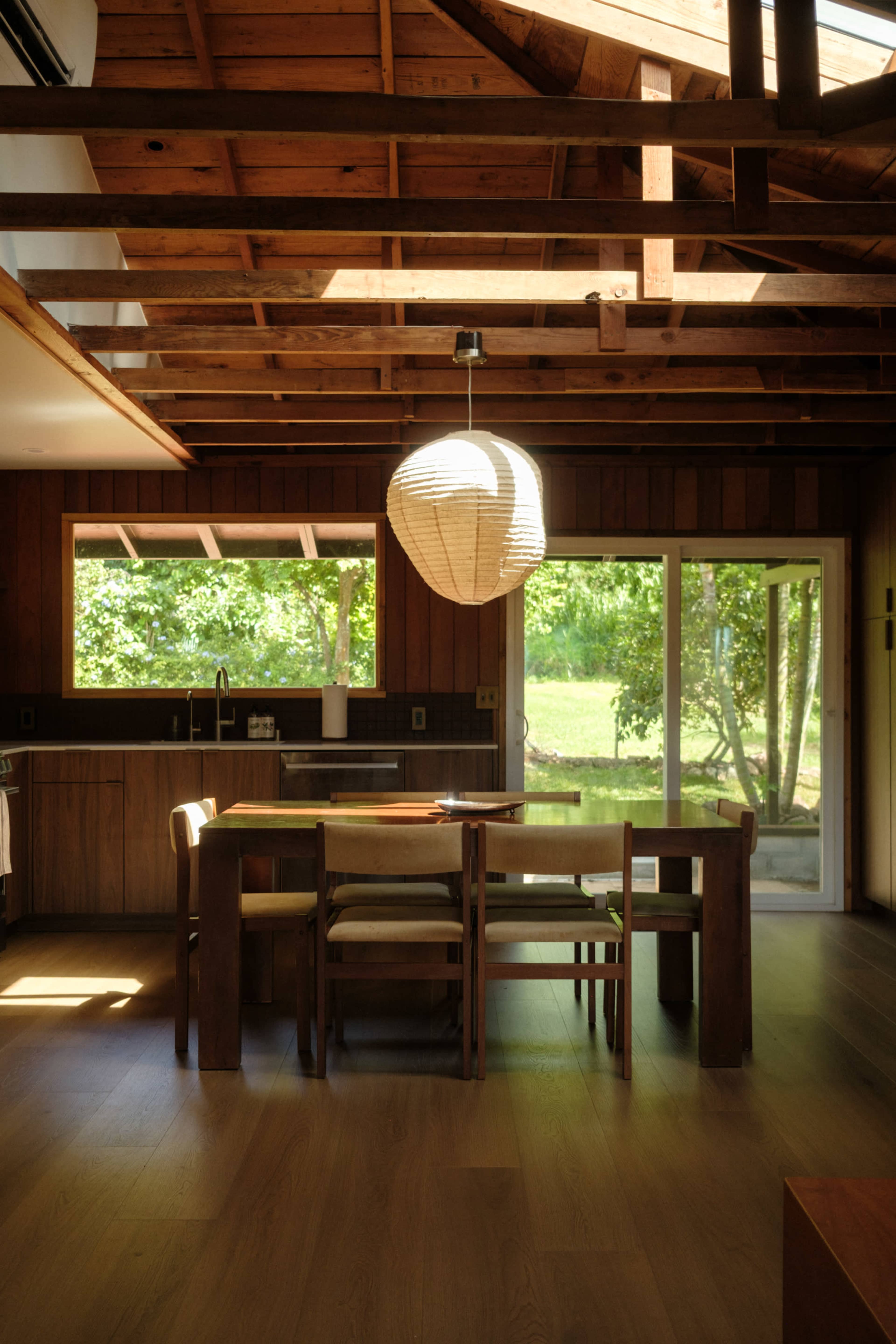 The image shows a modern kitchen-dining area with wooden beams, a table set with chairs, and large windows overlooking greenery.