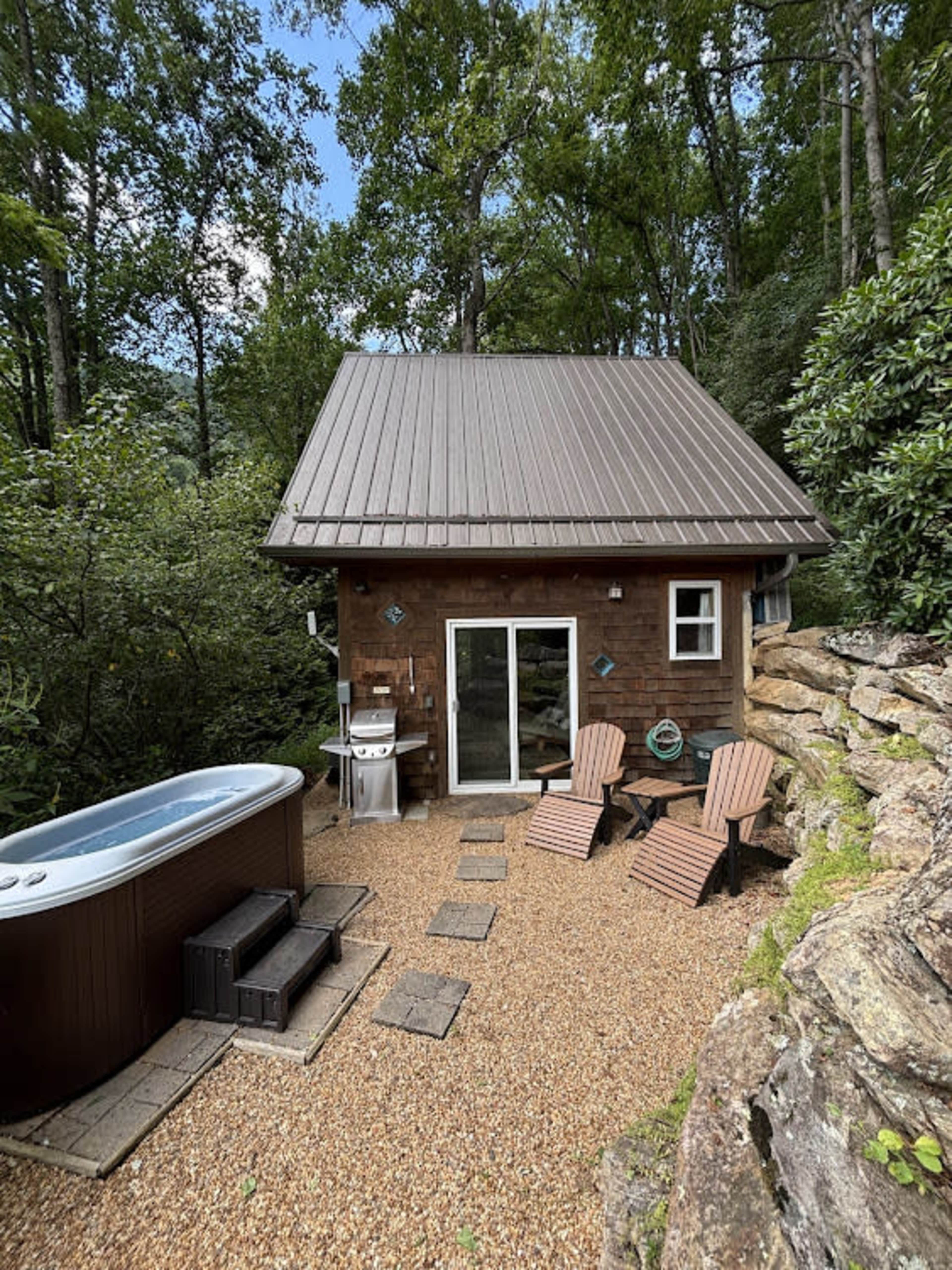 A small wooden cabin with a metal roof is situated among trees, featuring a hot tub, outdoor chairs, and a grill on a gravel patio.