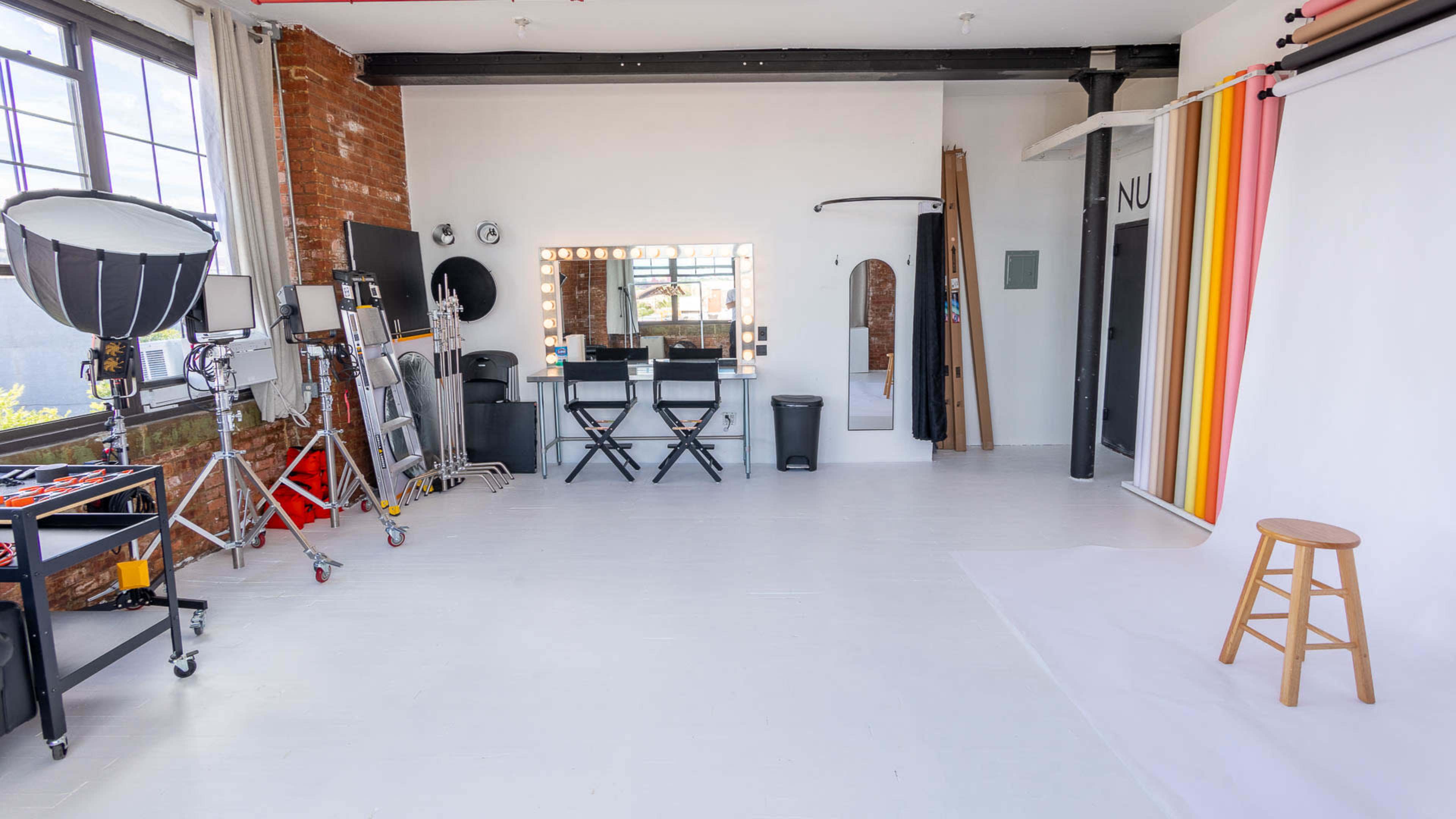 A spacious studio with a photography backdrop, lighting equipment, a makeup station with chairs, and a wooden stool positioned in the center.